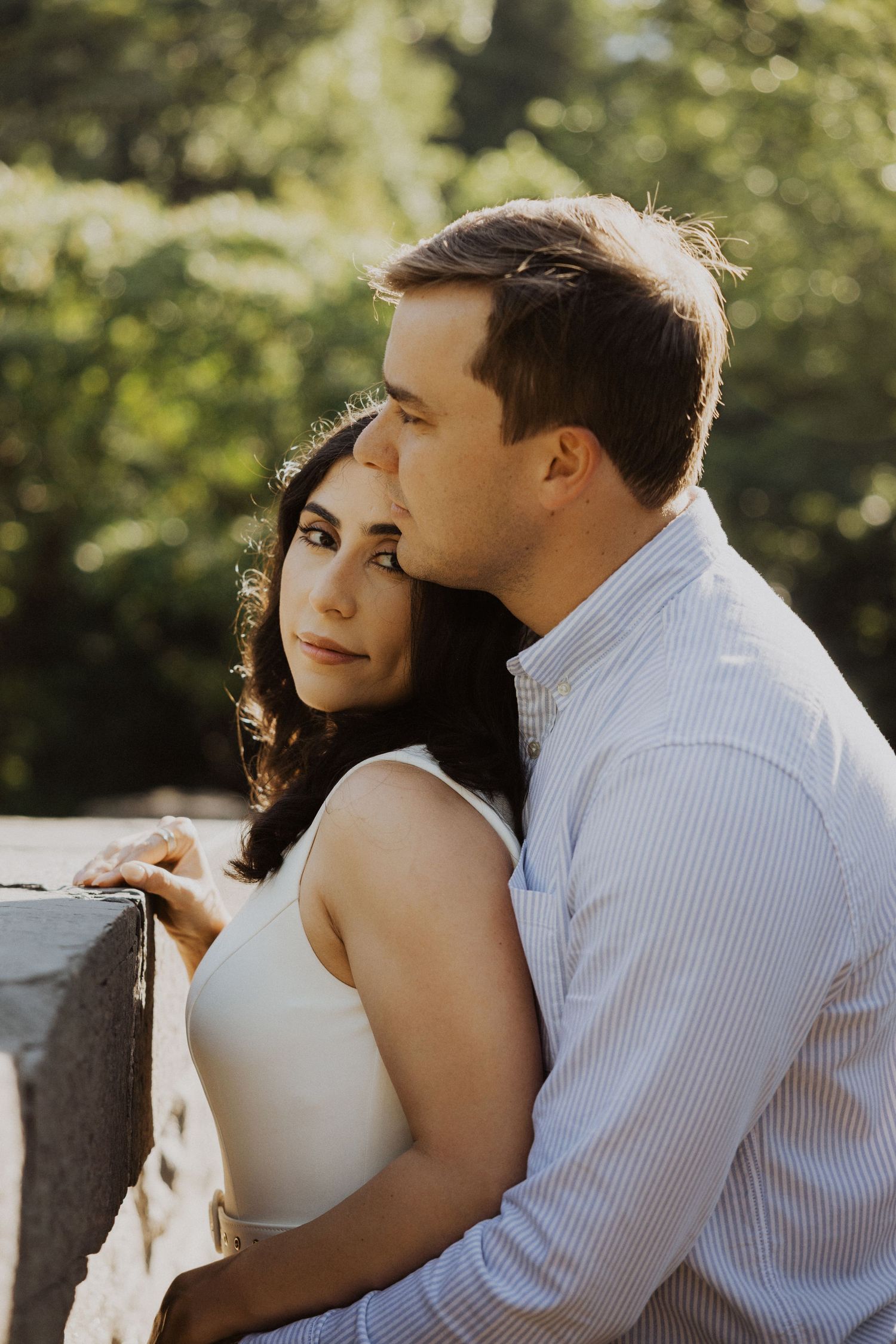 A couple shares a tender moment outdoors, the man embracing the woman from behind near a stone wall with lush green trees.