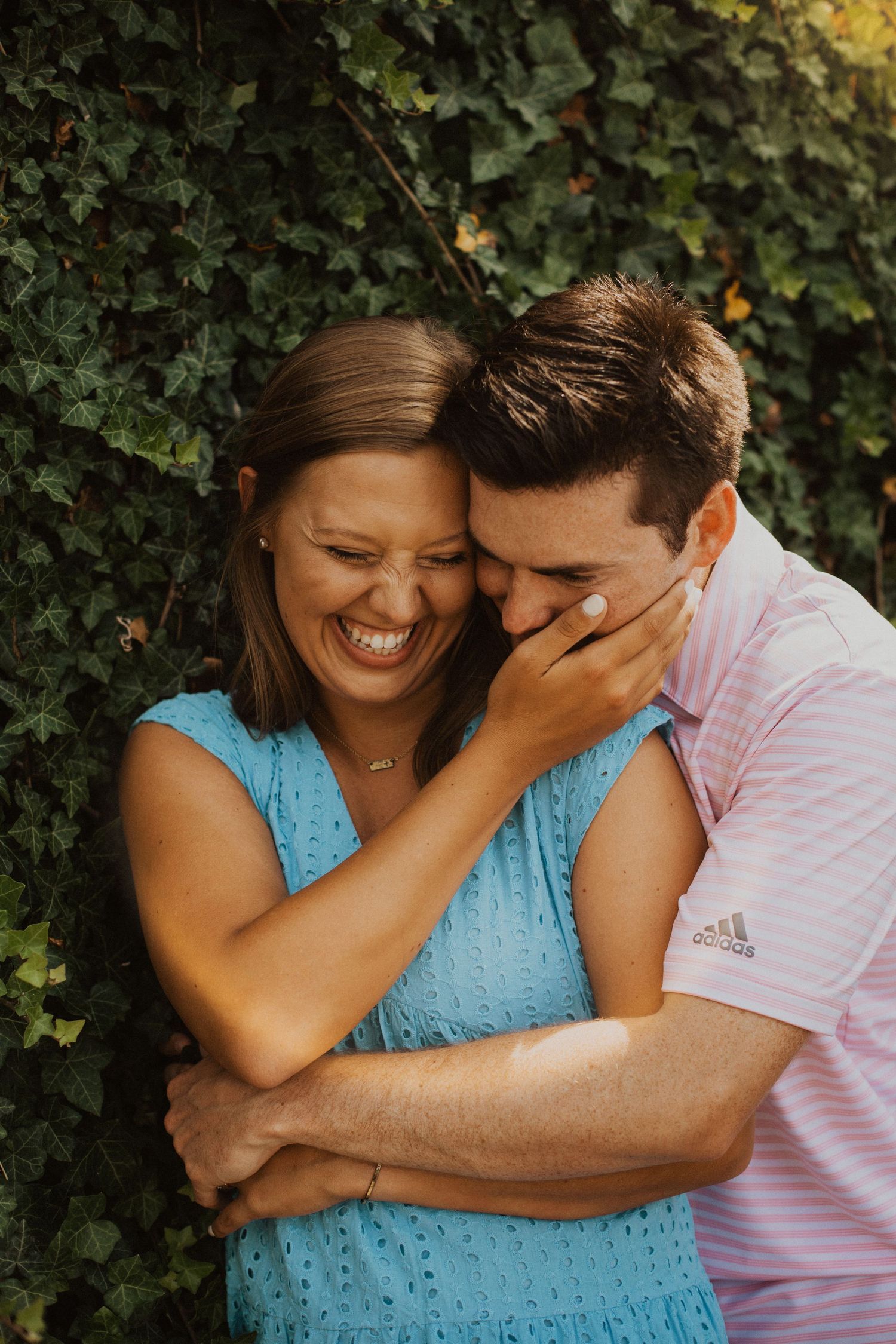 Couple laughs together against ivy-covered wall during engagement session.