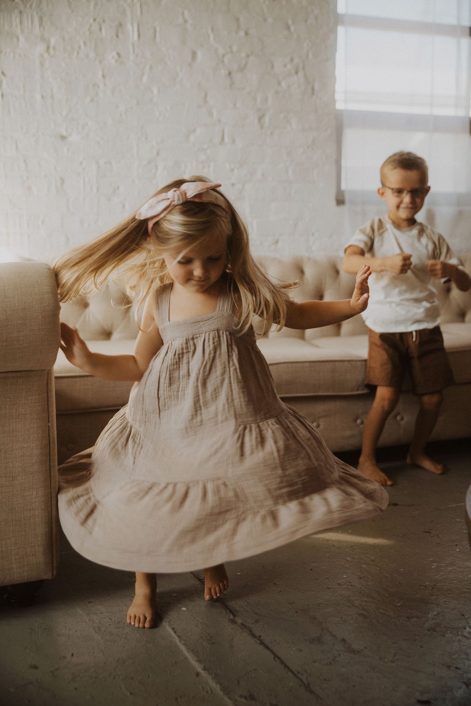 Young girl in beige tiered dress twirling barefoot indoors while a boy watches from a tufted sofa.