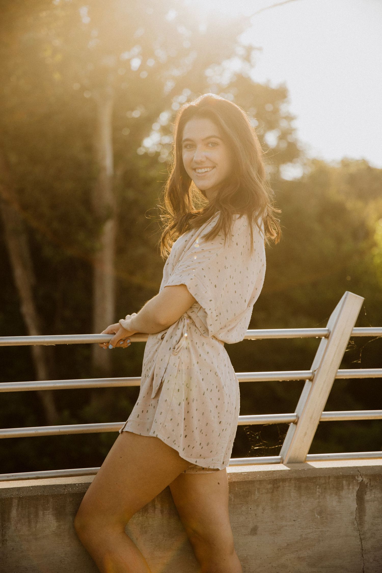 Young woman in floral romper smiling on bridge railing at golden hour sunset with trees in background.