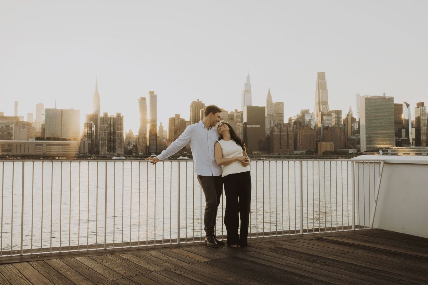 Couple embracing on a wooden deck with the New York City skyline at sunset in the background.