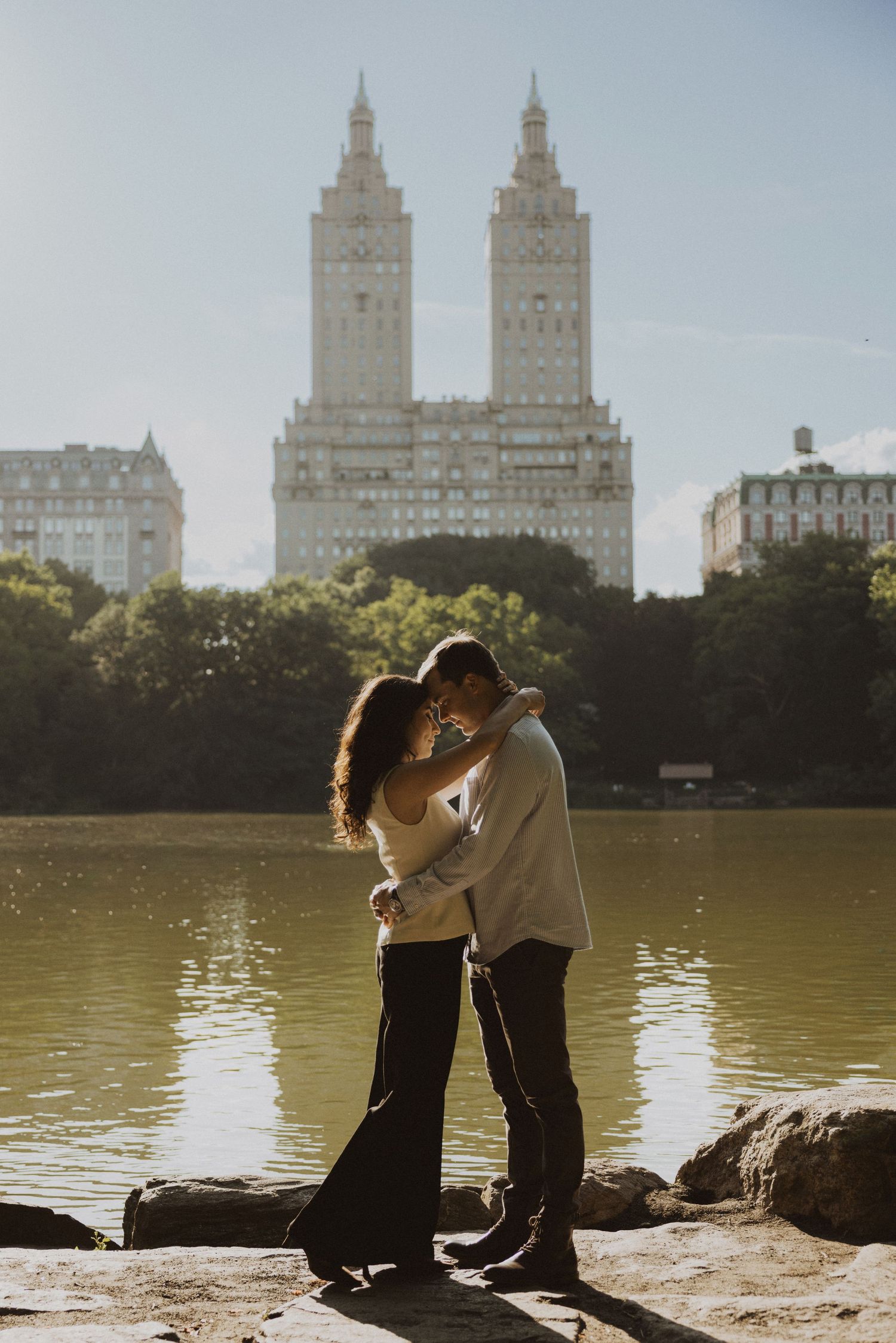 Couple embracing romantically by Central Park lake with iconic San Remo building twin towers in background, New York City.