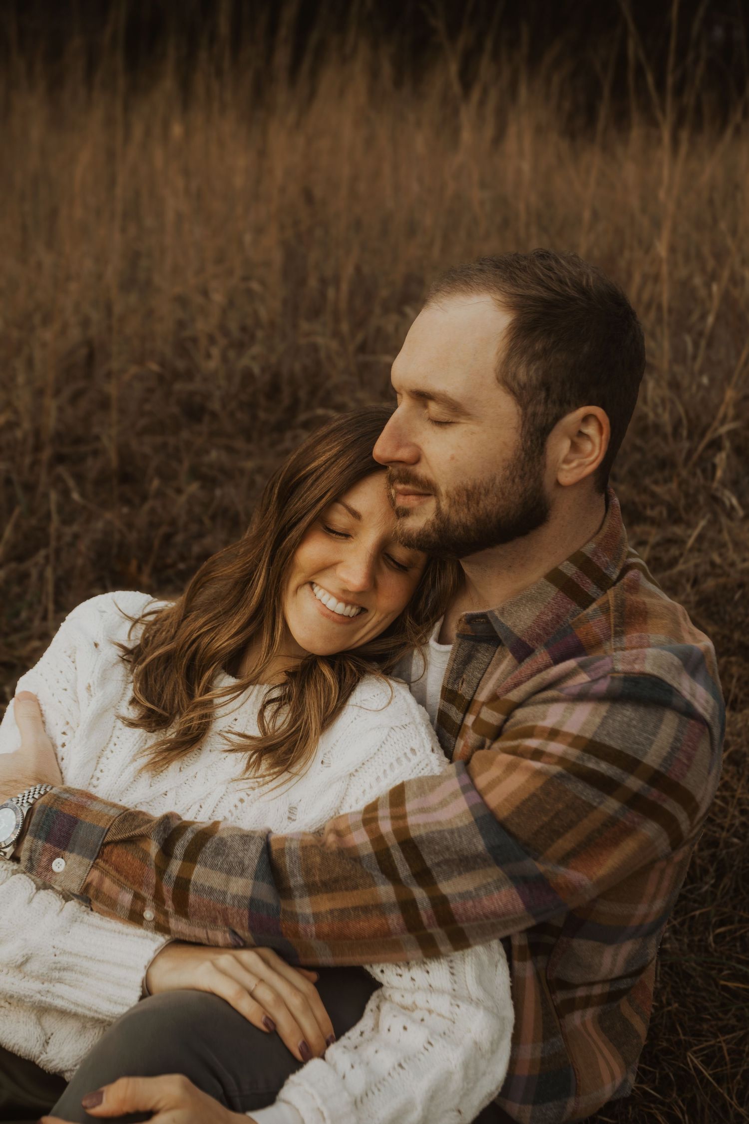 A couple sits in a golden field, sharing a tender embrace as the man kisses the smiling woman on the cheek.