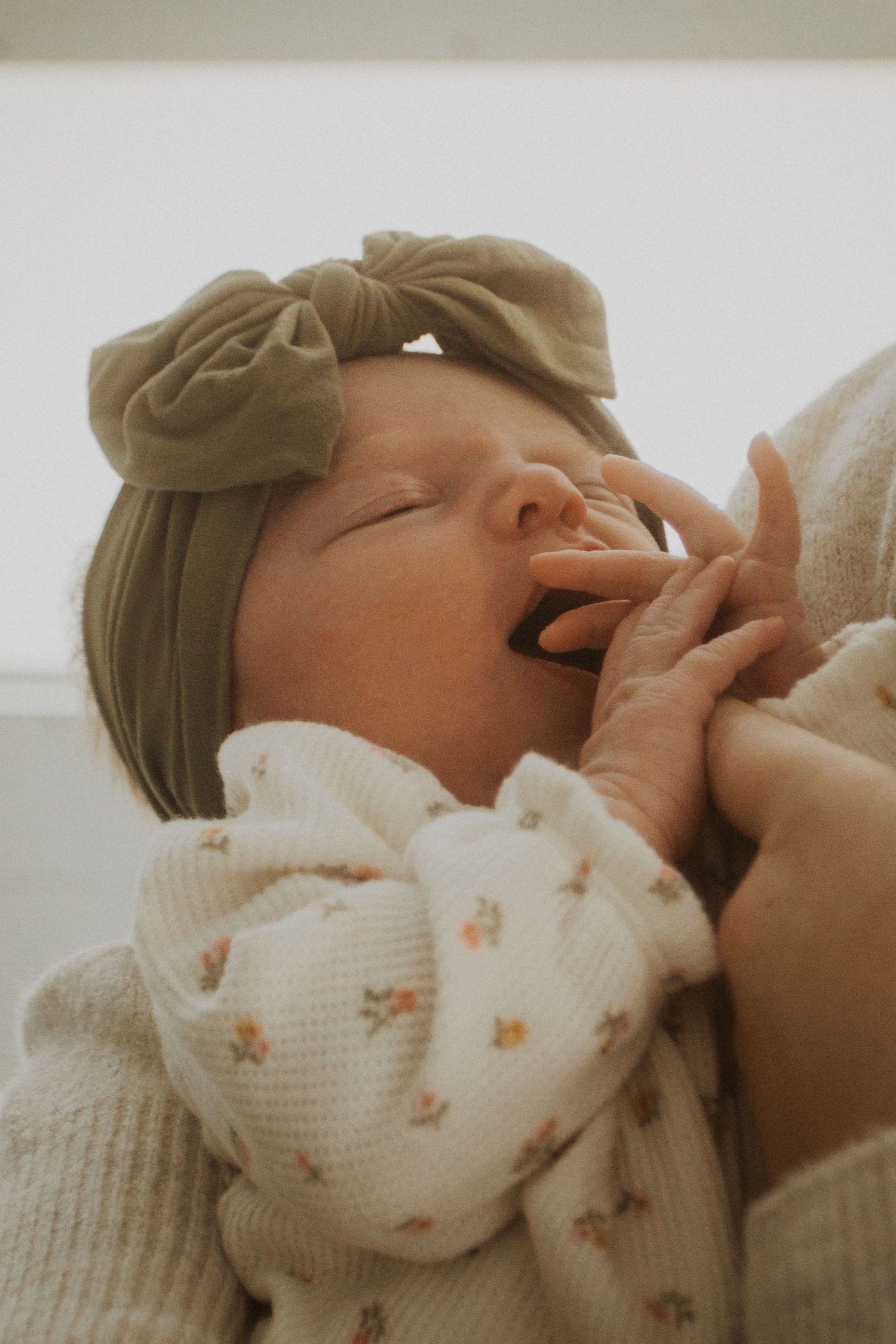 Newborn baby yawning with olive green bow headband, wearing floral onesie, cradled in mother's arms.