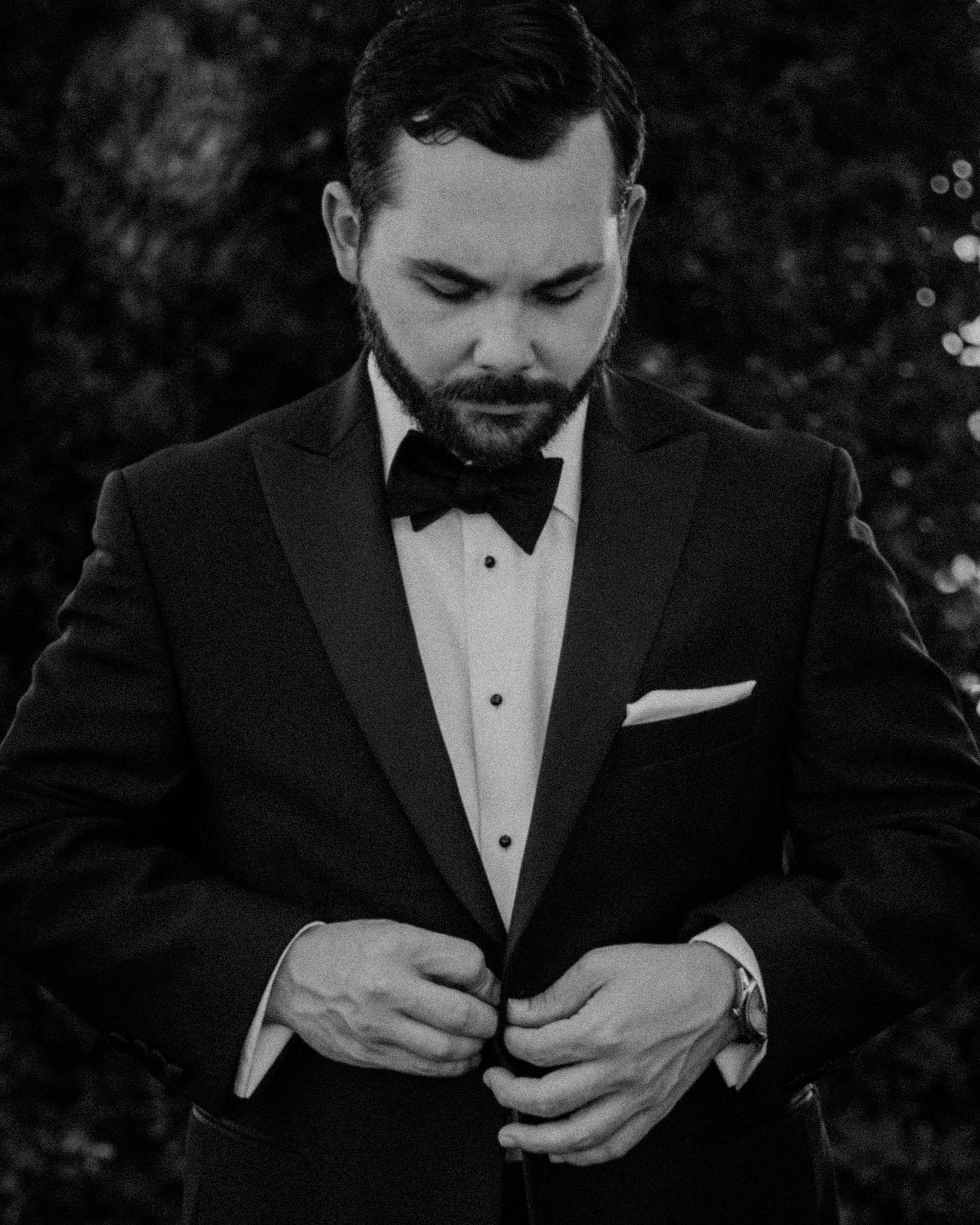A groom in a classic black tuxedo adjusts his bowtie in a black and white portrait series.