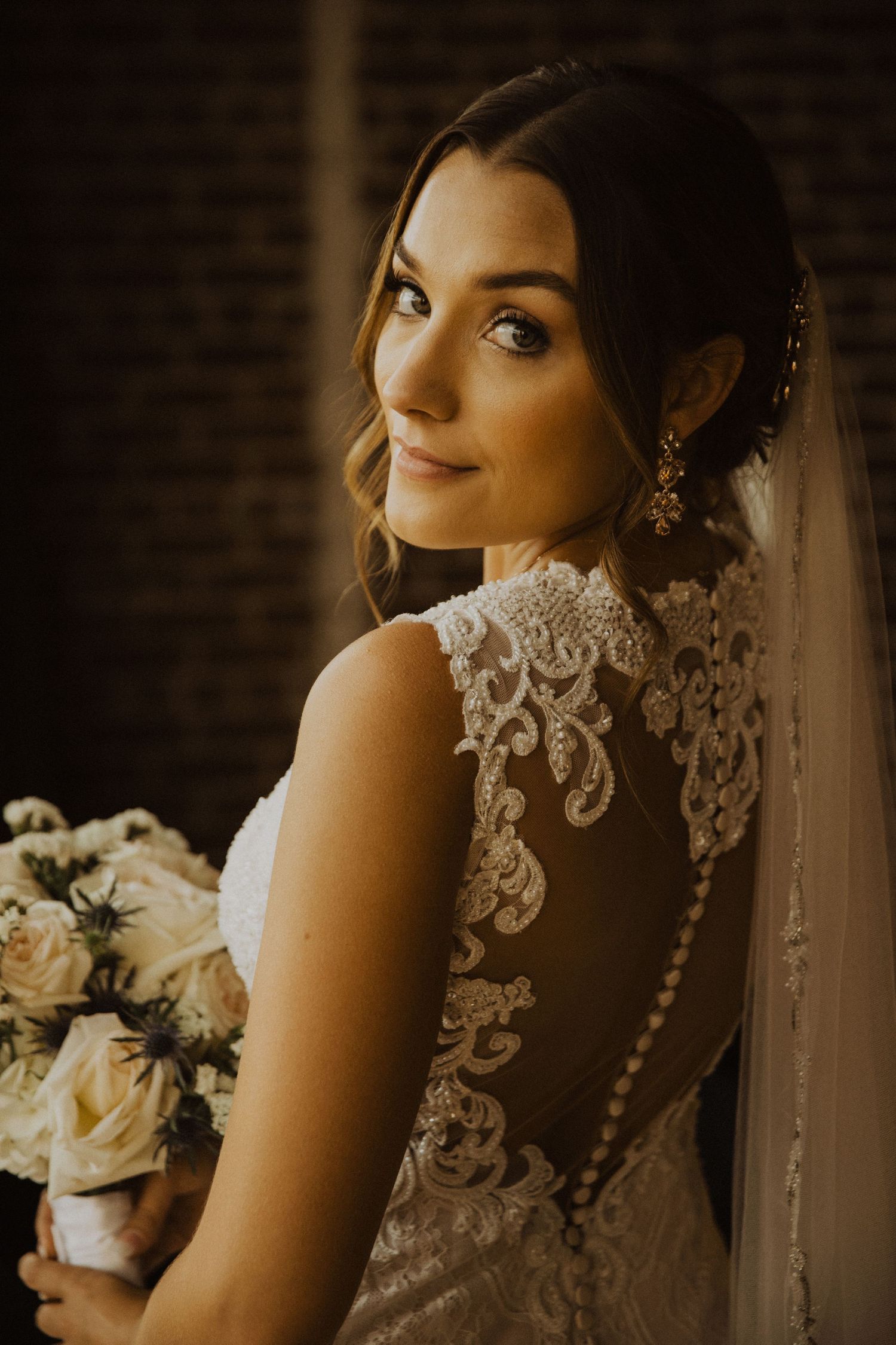 Bride with floral bouquet poses in detailed lace gown near window with romantic lighting.