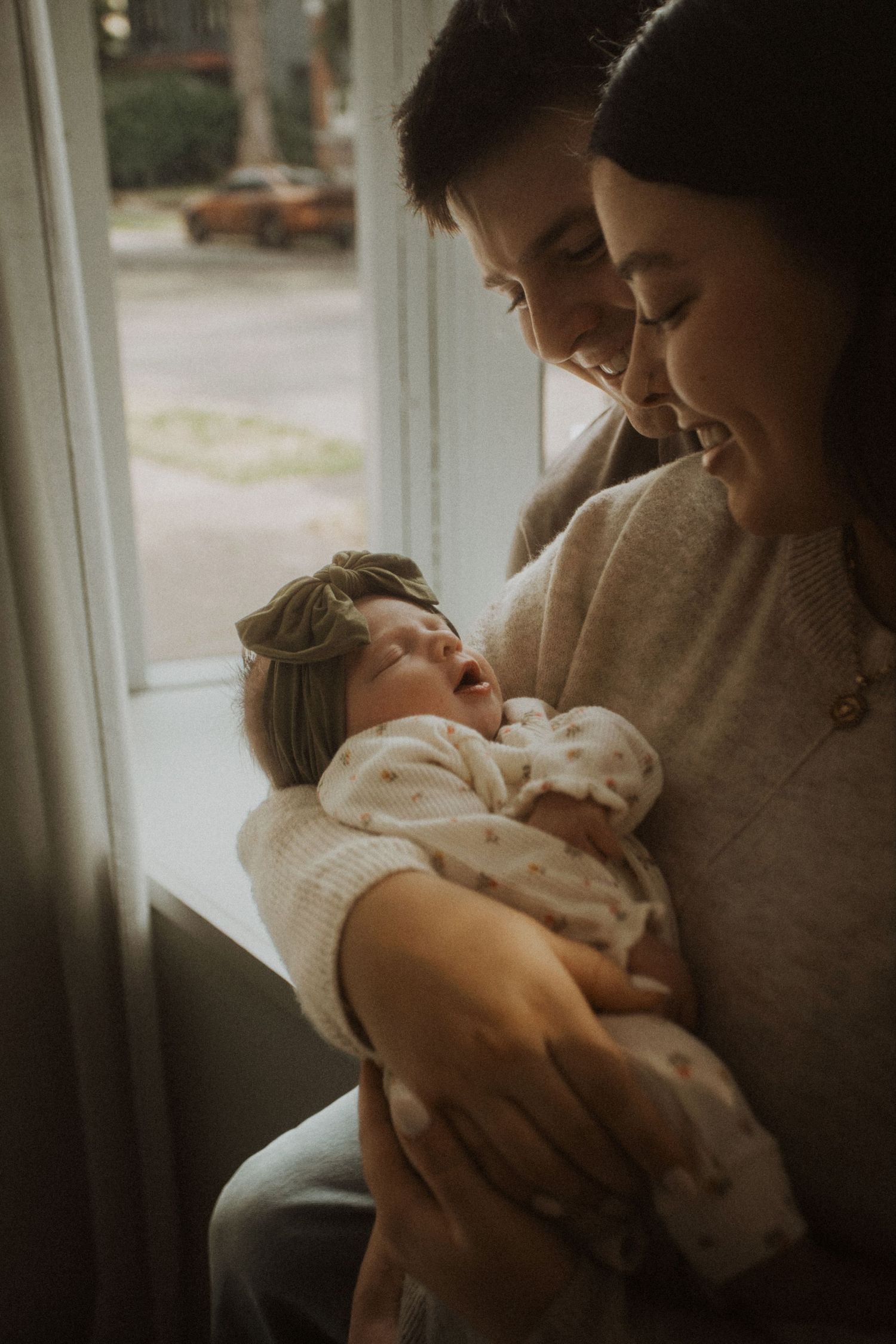 Two smiling parents lovingly hold their newborn baby girl wearing a green bow headband near a bright window.