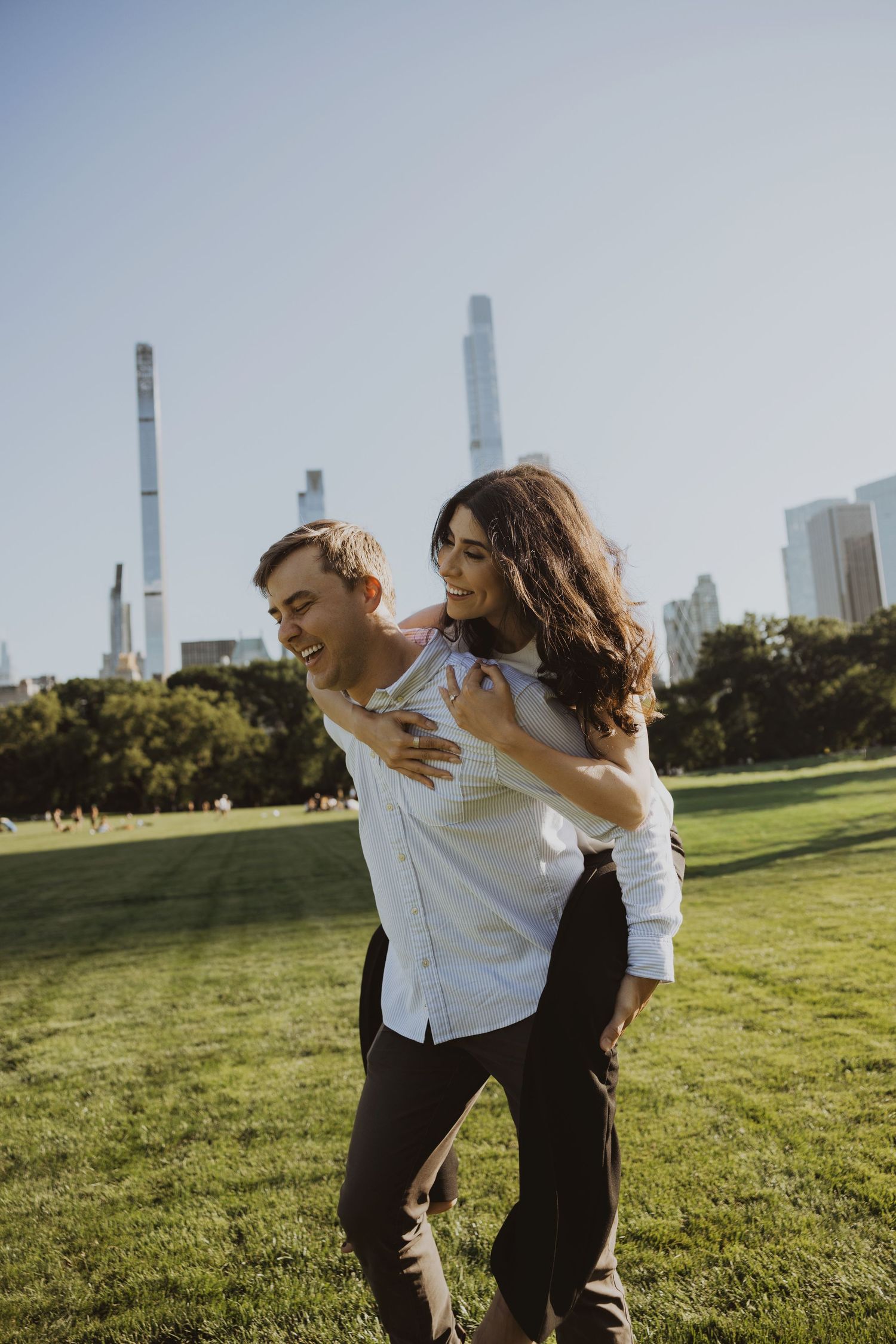 A happy couple enjoys a piggyback ride in Central Park with NYC skyline in the background on a sunny day.