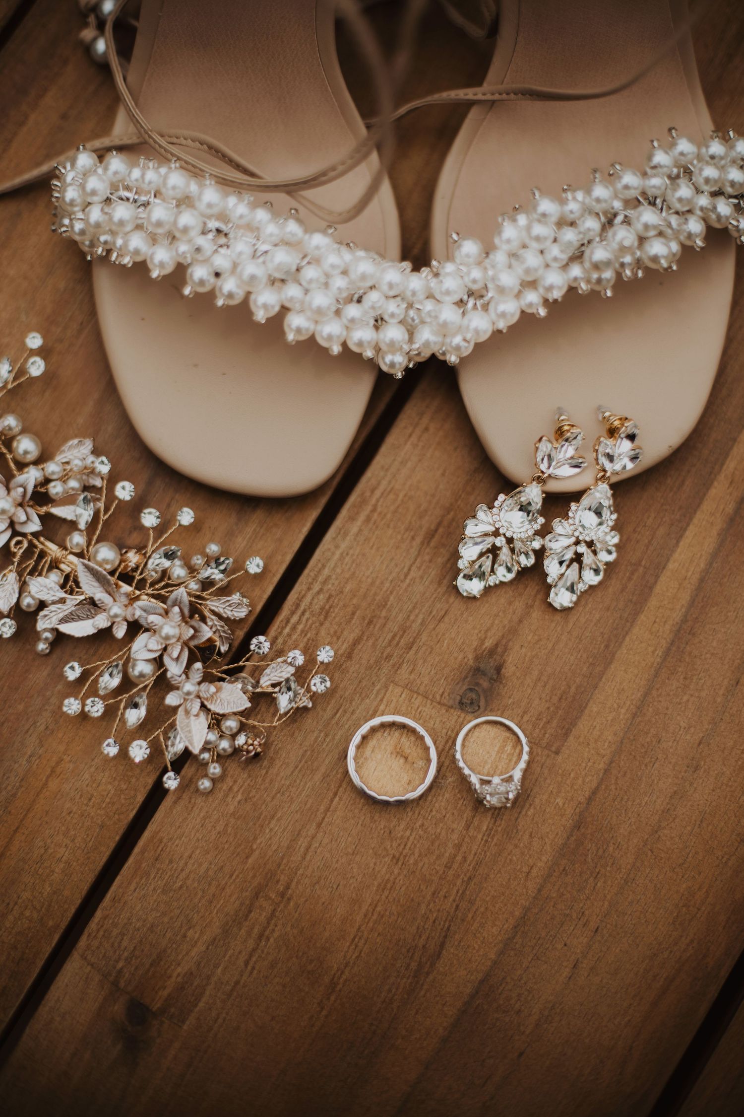 Wedding accessories including pearl headpiece, rings, and crystal jewelry arranged on rustic wooden surface.