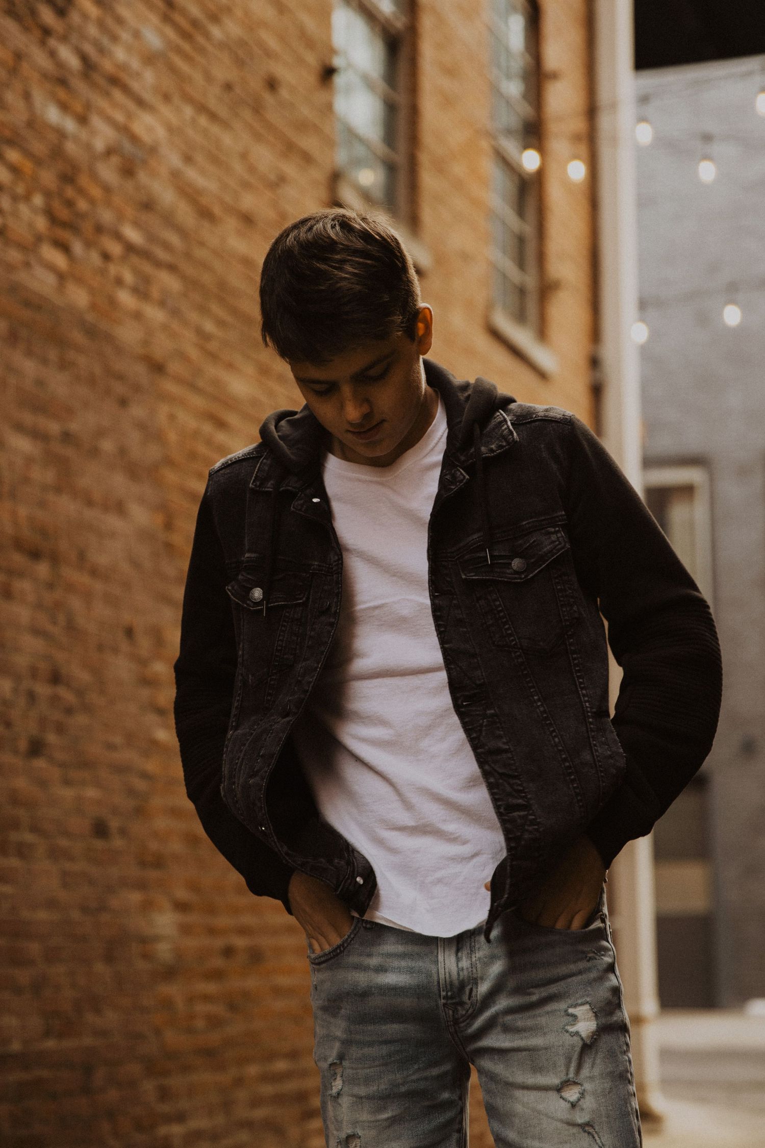 Young man in black denim jacket and white tee poses in urban alley with brick wall backdrop.