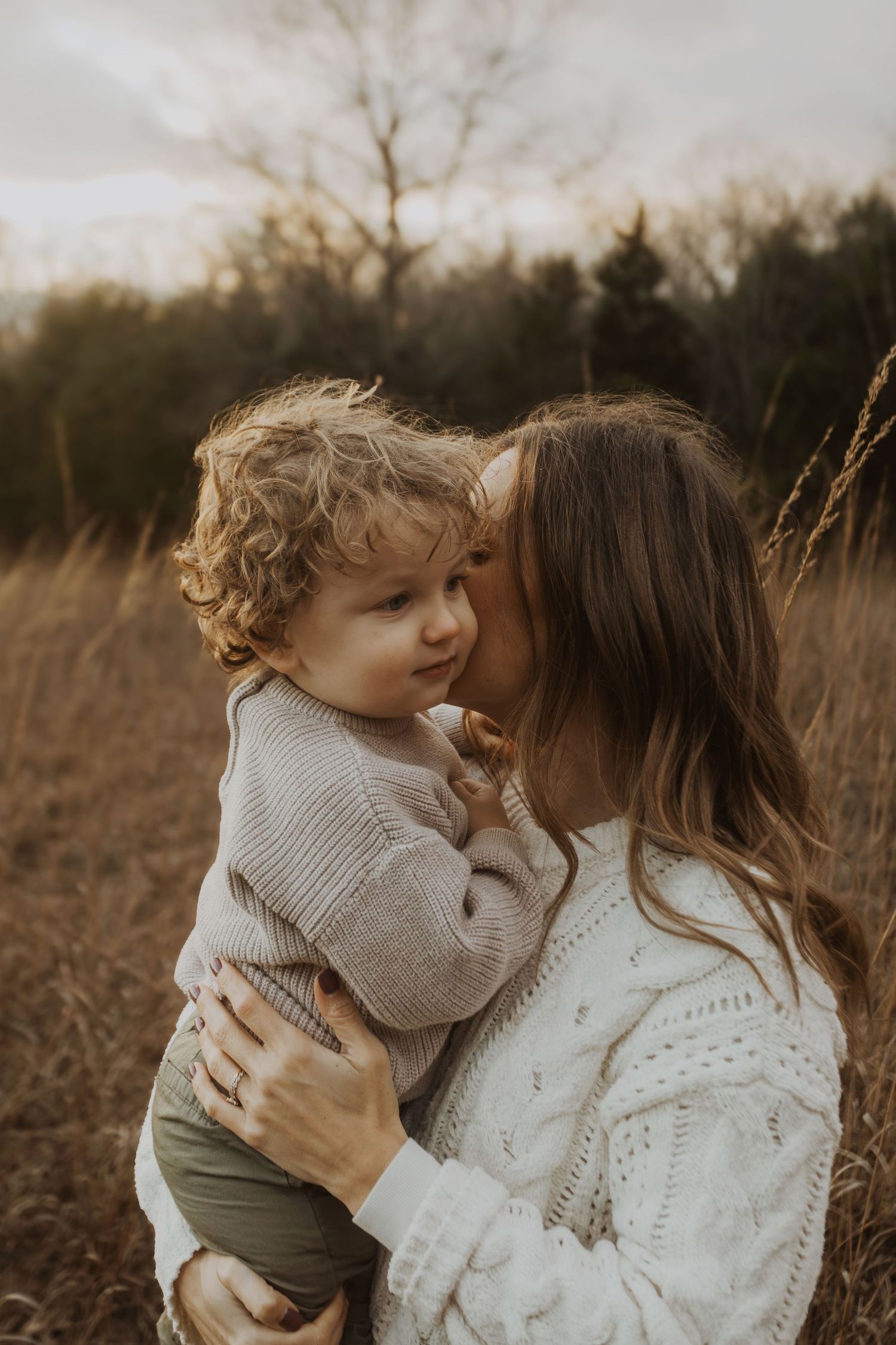A mother tenderly kisses her curly-haired toddler in a golden autumn field at dusk.