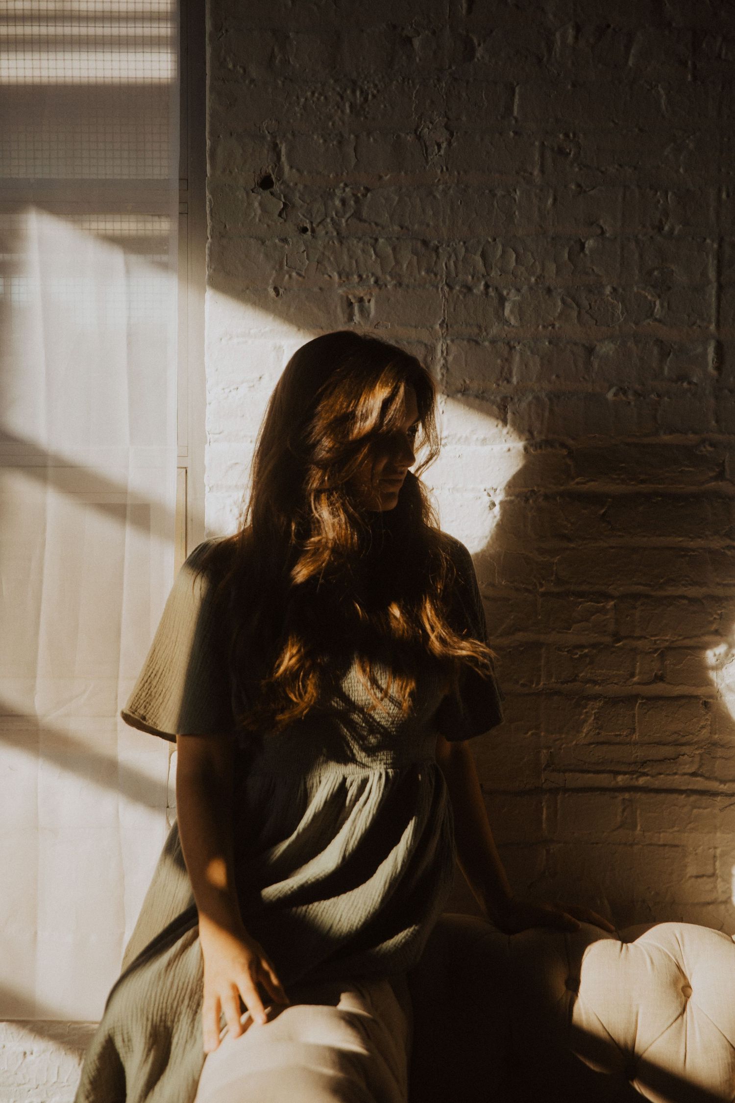 A woman with long wavy hair sits in warm golden sunlight by a brick wall, wearing a sage green dress.