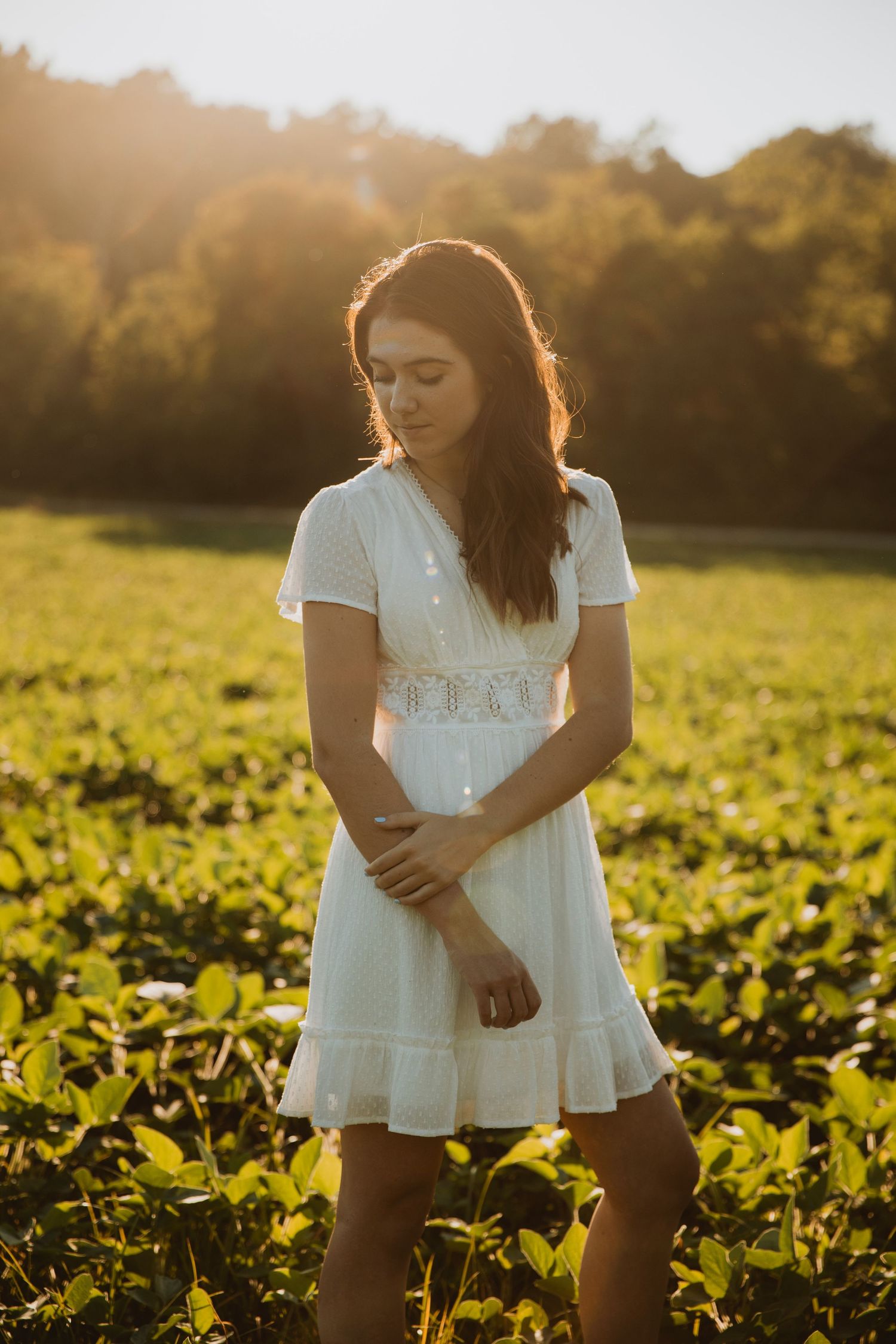 Young woman in white lace dress standing in green field during golden hour sunset.
