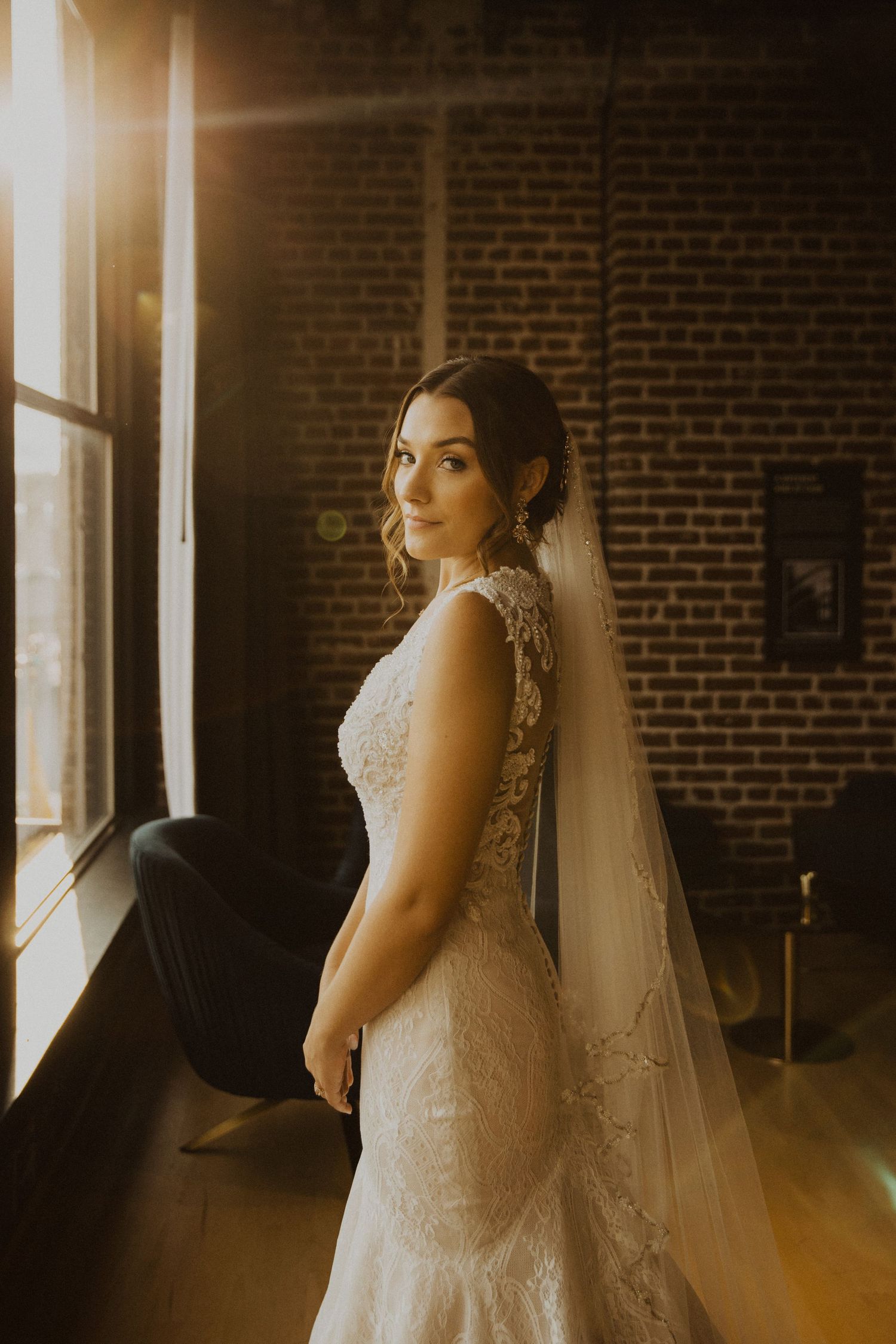 Bride in elegant lace wedding dress poses near window with exposed brick wall in moody lighting.
