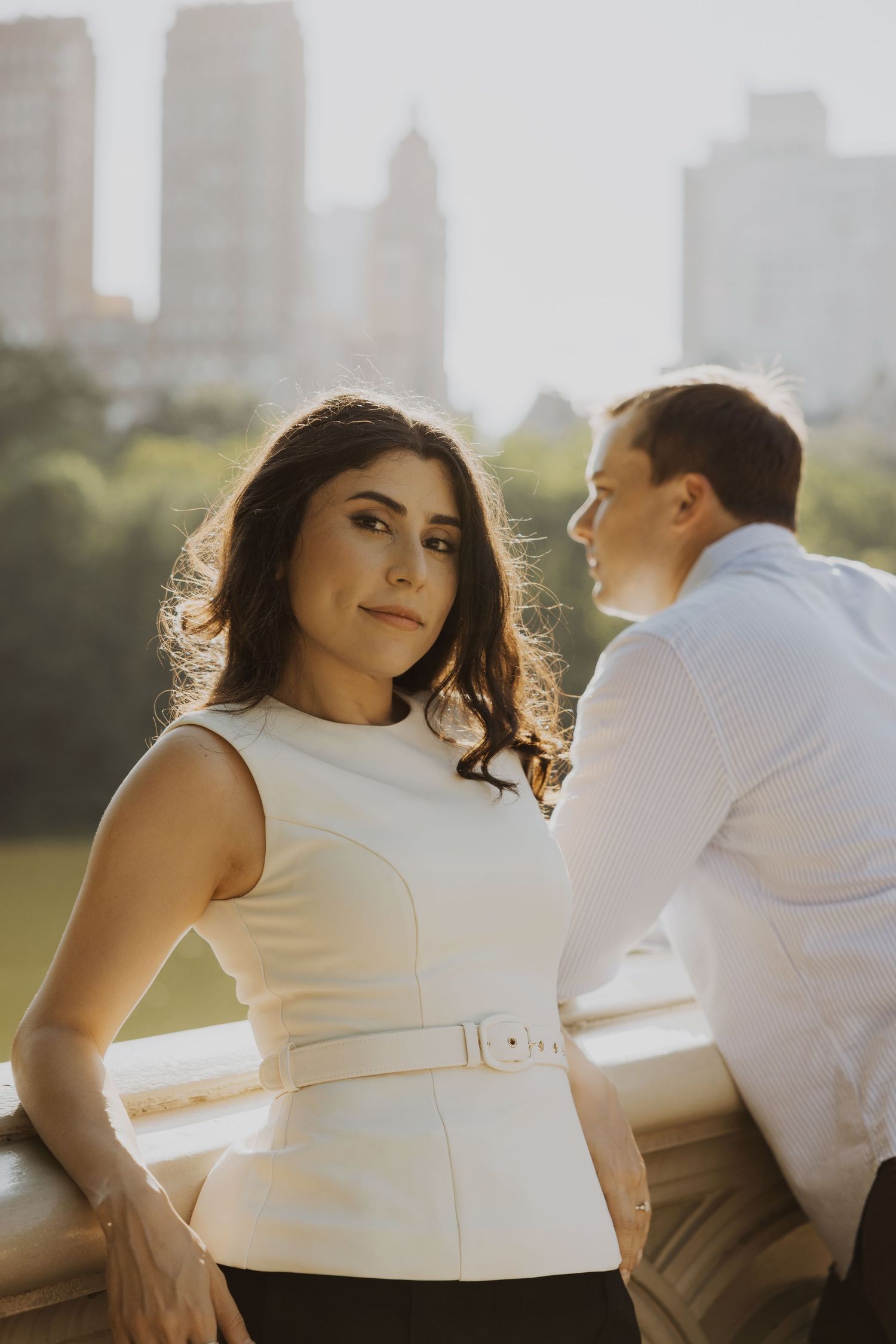 A woman in a white sleeveless dress poses on a bridge in Central Park, NYC, with a man and city skyline behind her.