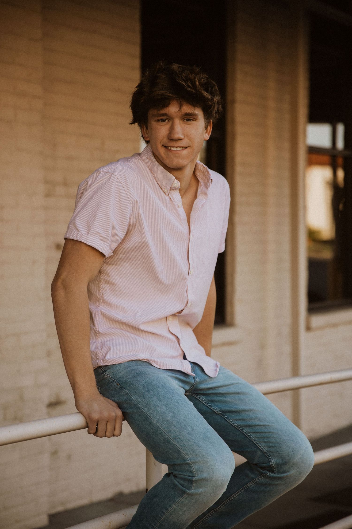 Young man in pink shirt and jeans smiling while leaning on a railing outside a brick building.