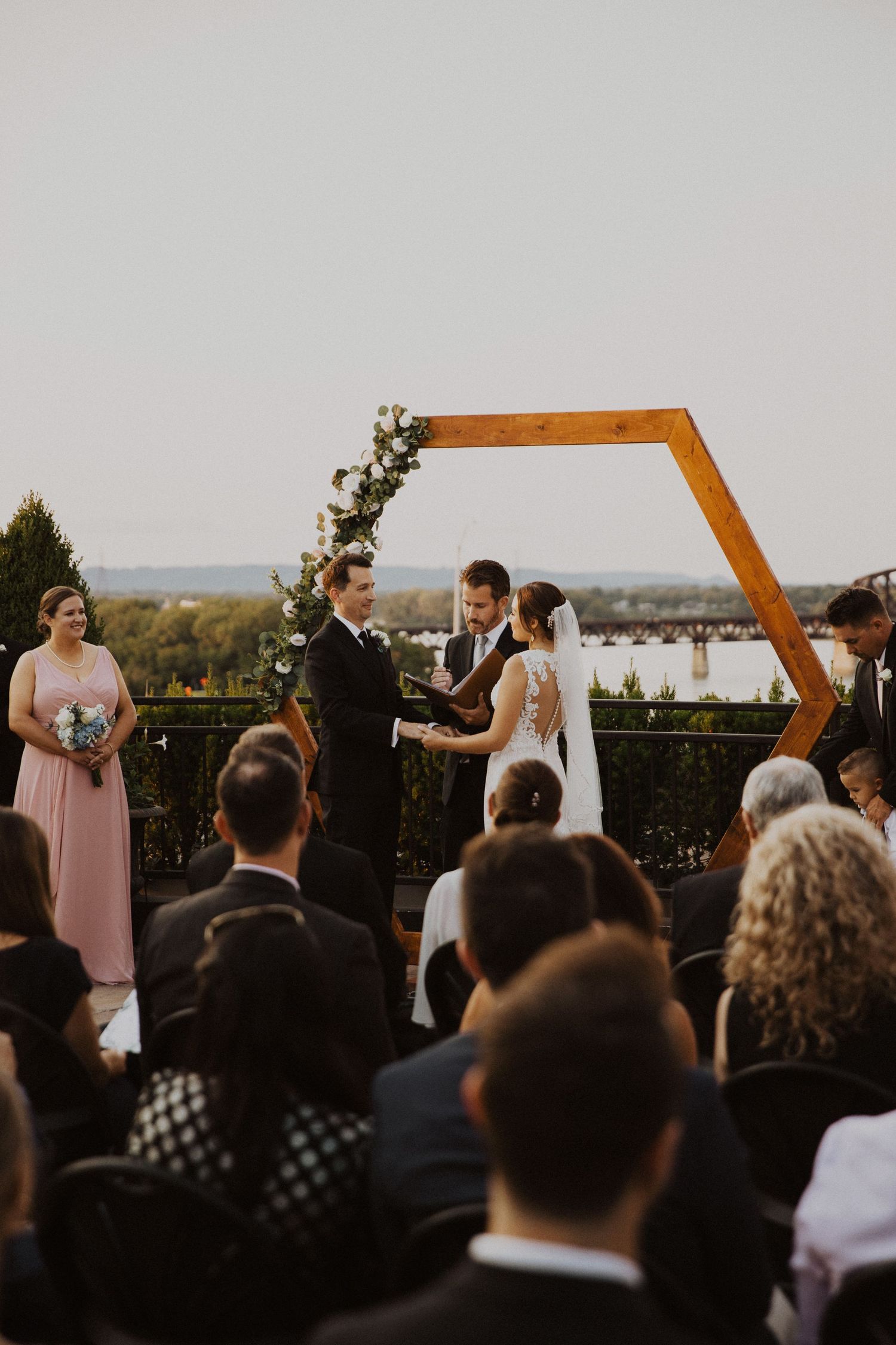 Outdoor wedding ceremony under wooden hexagonal arch with guests seated on rooftop.