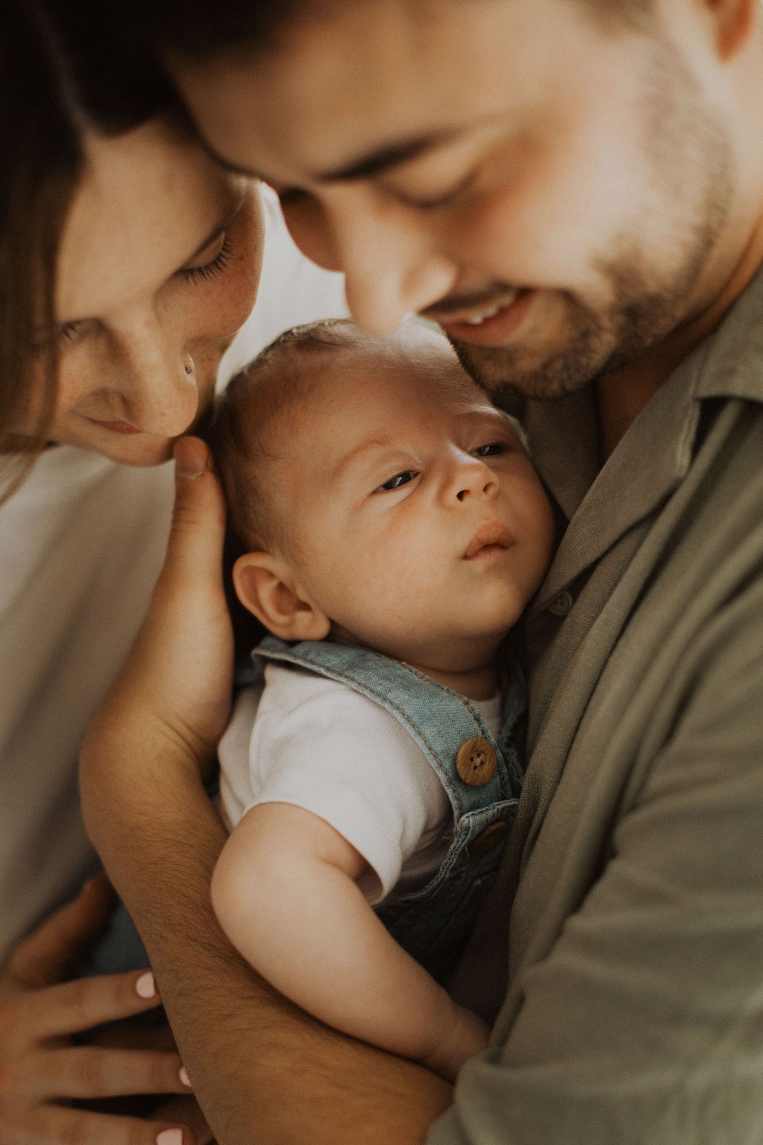 Two loving parents holding their newborn baby close, sharing a tender family moment in warm natural light.