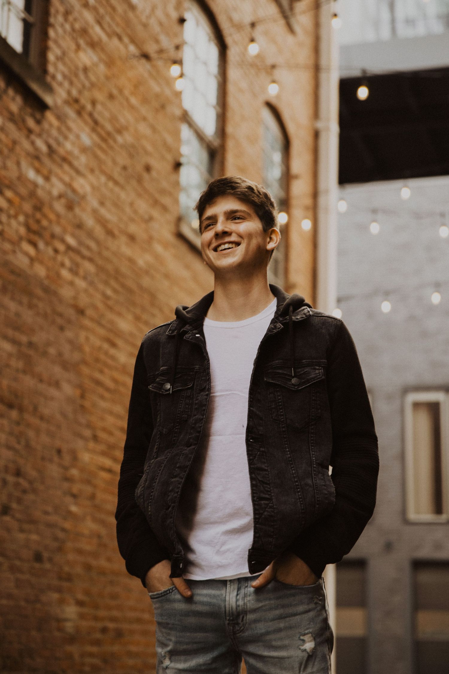 Smiling young man in black denim jacket and white tee, standing outdoors near brick wall with string lights.