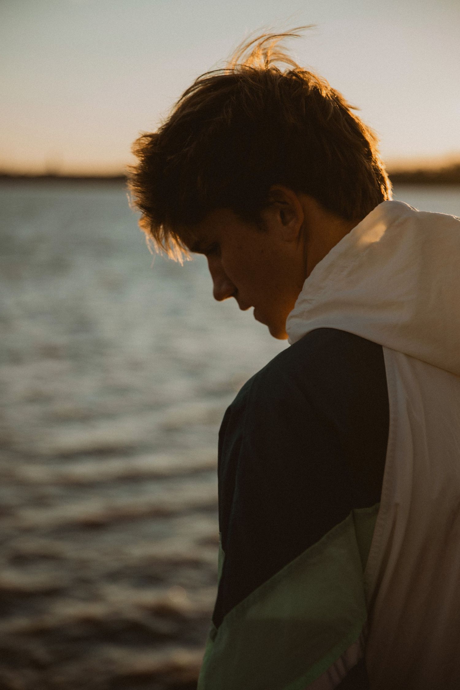 Young man in a white jacket gazing down by a serene lake at golden hour sunset.