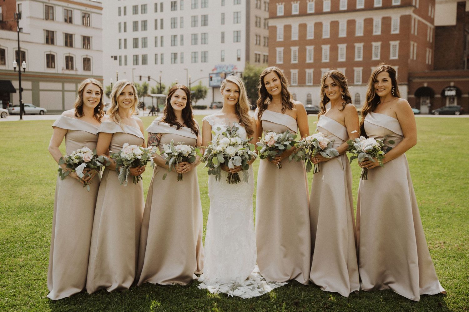 Bridal party in neutral dresses poses with bouquets outdoors.