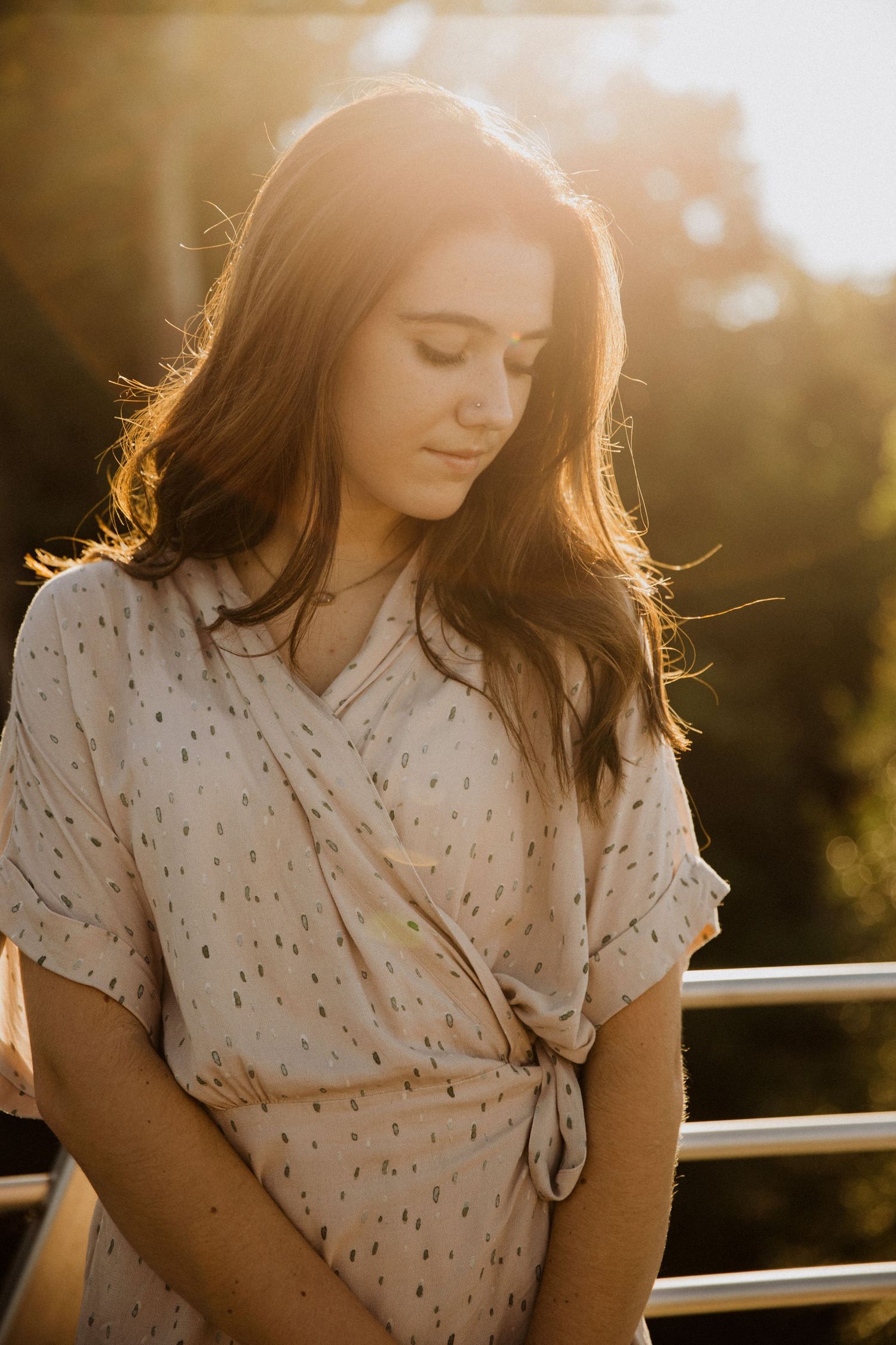 Young woman in a floral wrap dress bathed in golden sunset light, eyes downcast, standing near metal railings outdoors.