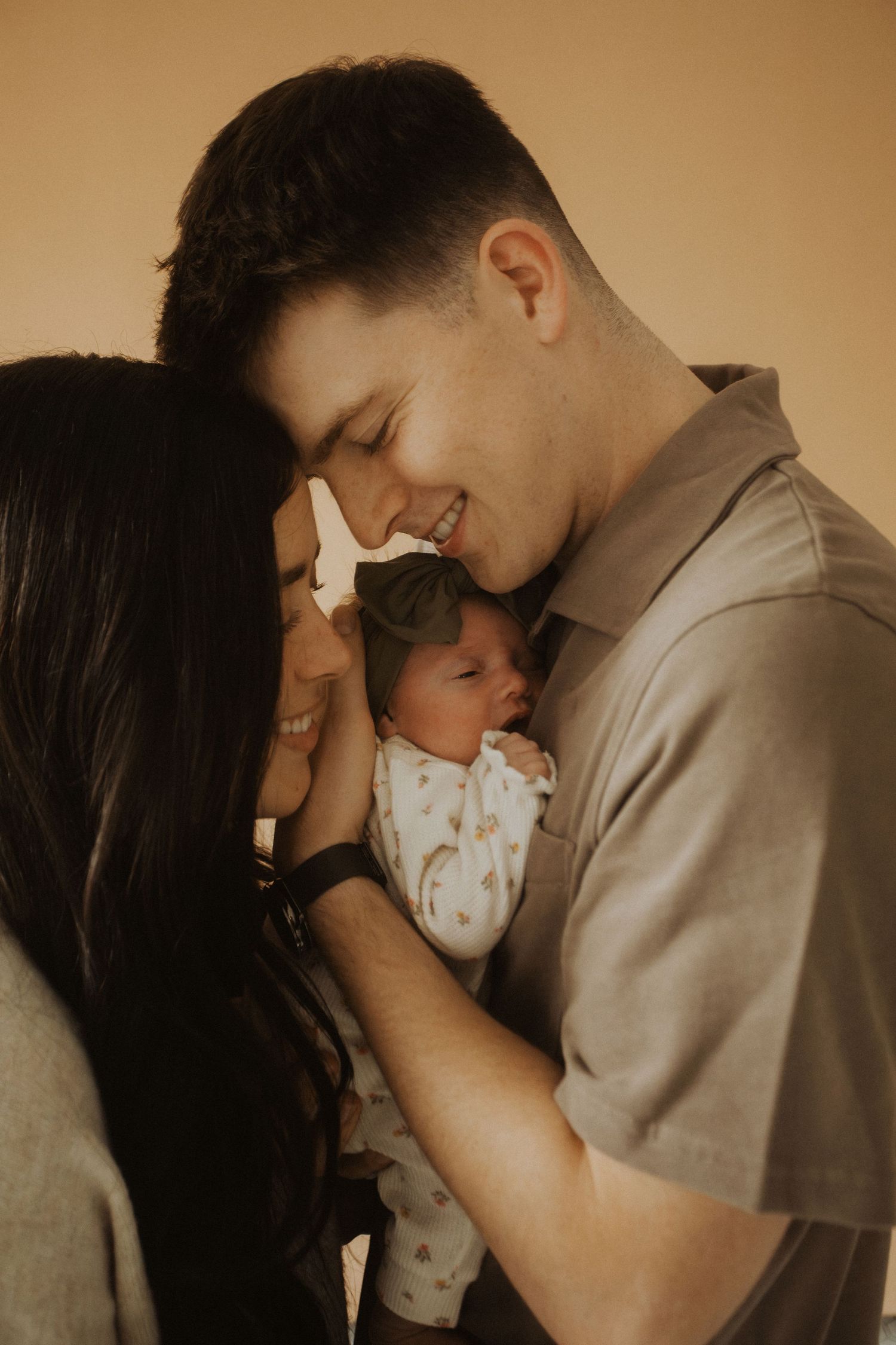 Happy couple smiling and holding their newborn baby together against a warm beige background.
