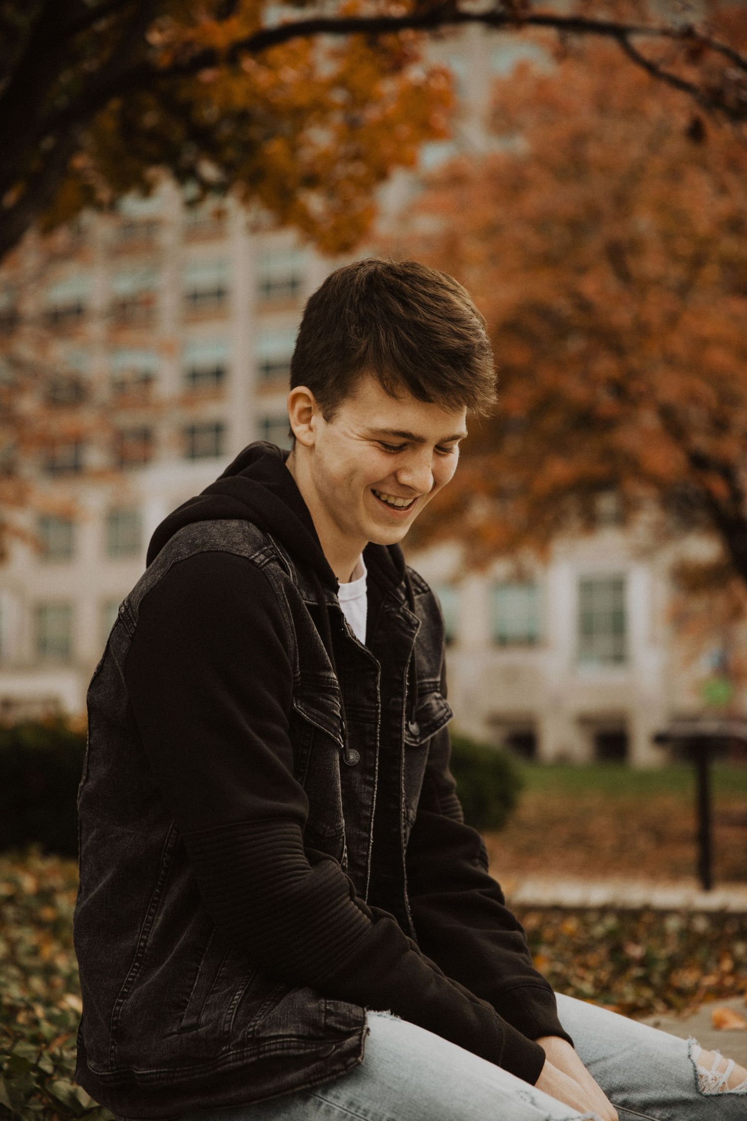 Young man smiling outdoors in autumn park, wearing black denim jacket over hoodie, seated among fall foliage.