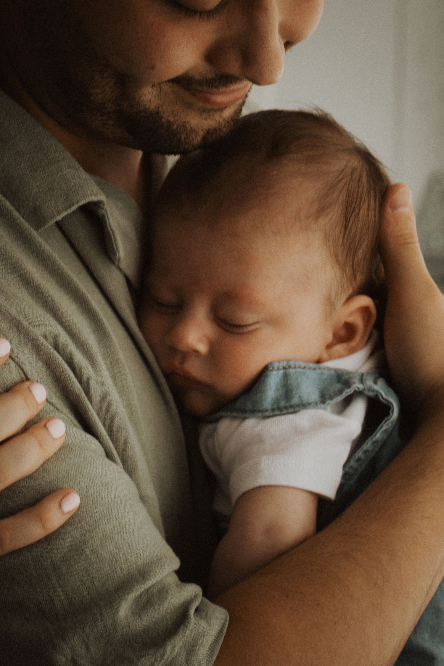 A bearded father tenderly holds his sleeping newborn baby dressed in denim overalls against his chest.