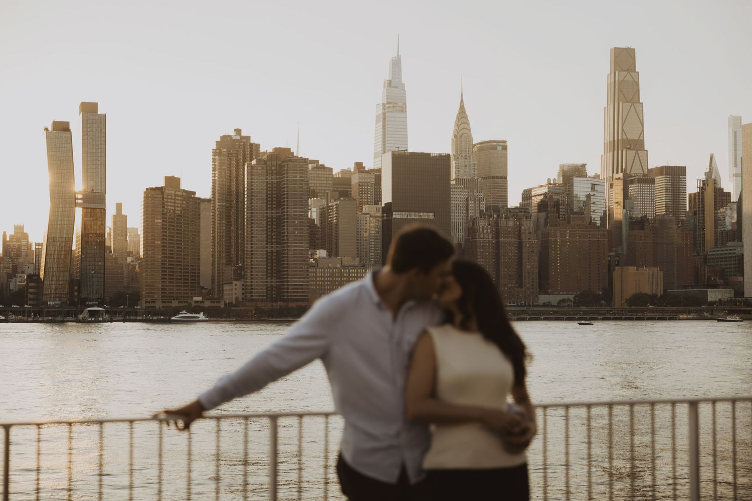 Couple sharing a romantic kiss with the stunning New York City skyline and river in the golden hour background.