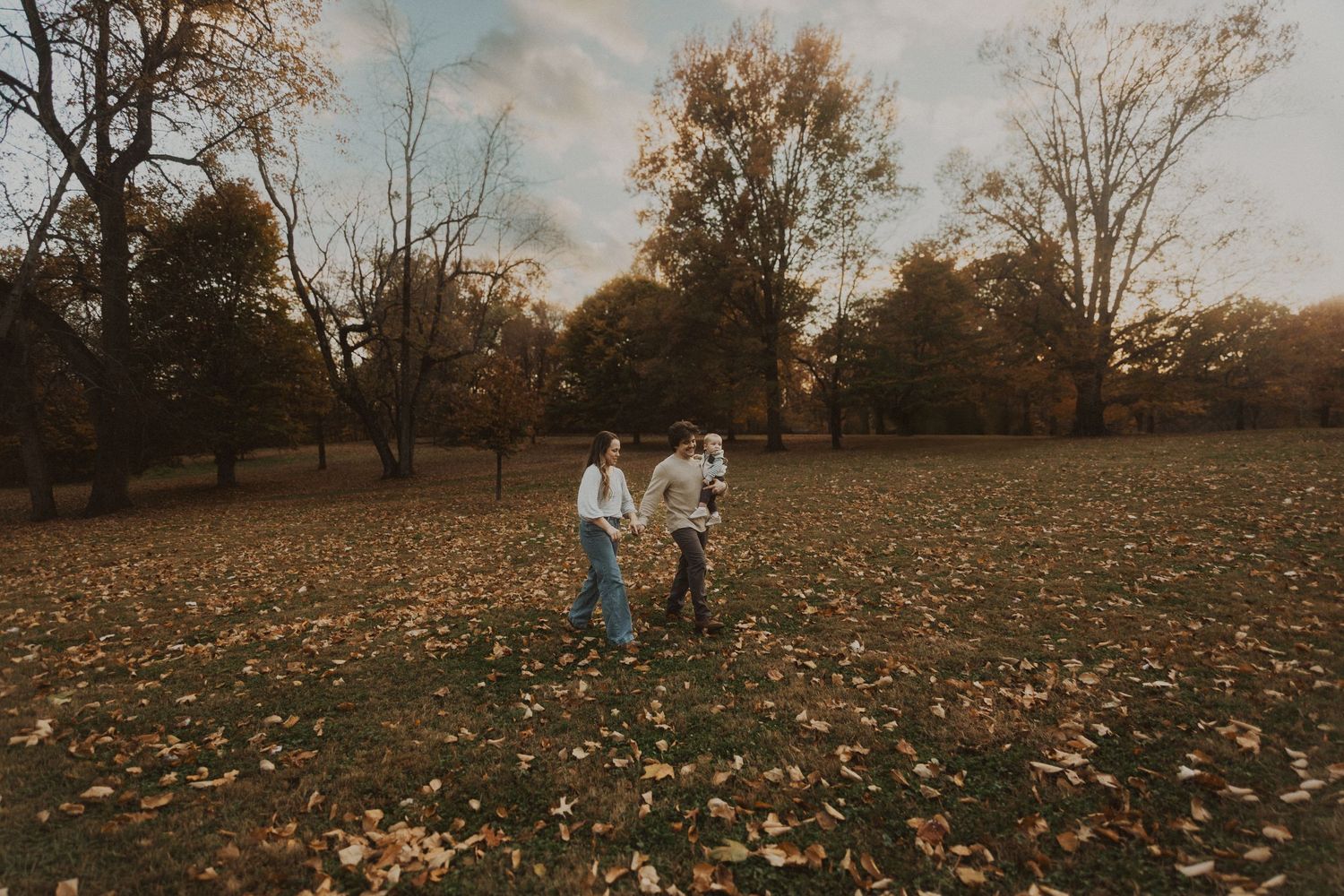 Family walking through a leaf-covered park during autumn, surrounded by golden trees in warm fall light.