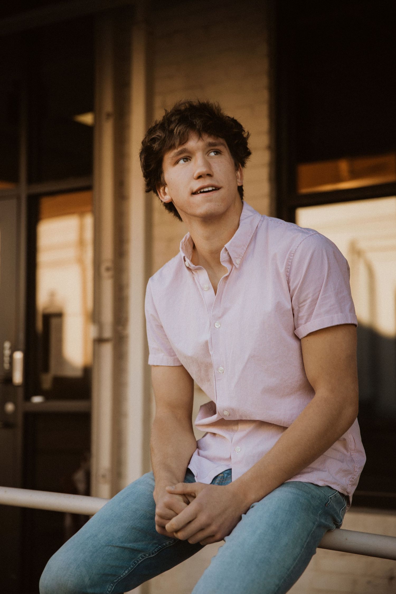 Young man with curly hair wearing a pink button-up shirt and jeans, sitting outdoors looking upward.