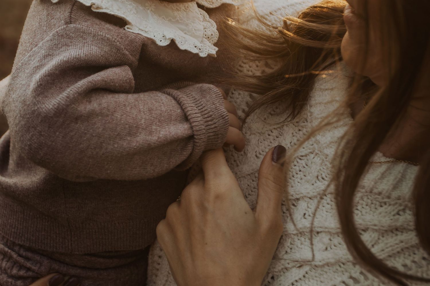 A mother in a white knit sweater tenderly holds her toddler dressed in a brown knit outfit in warm light.