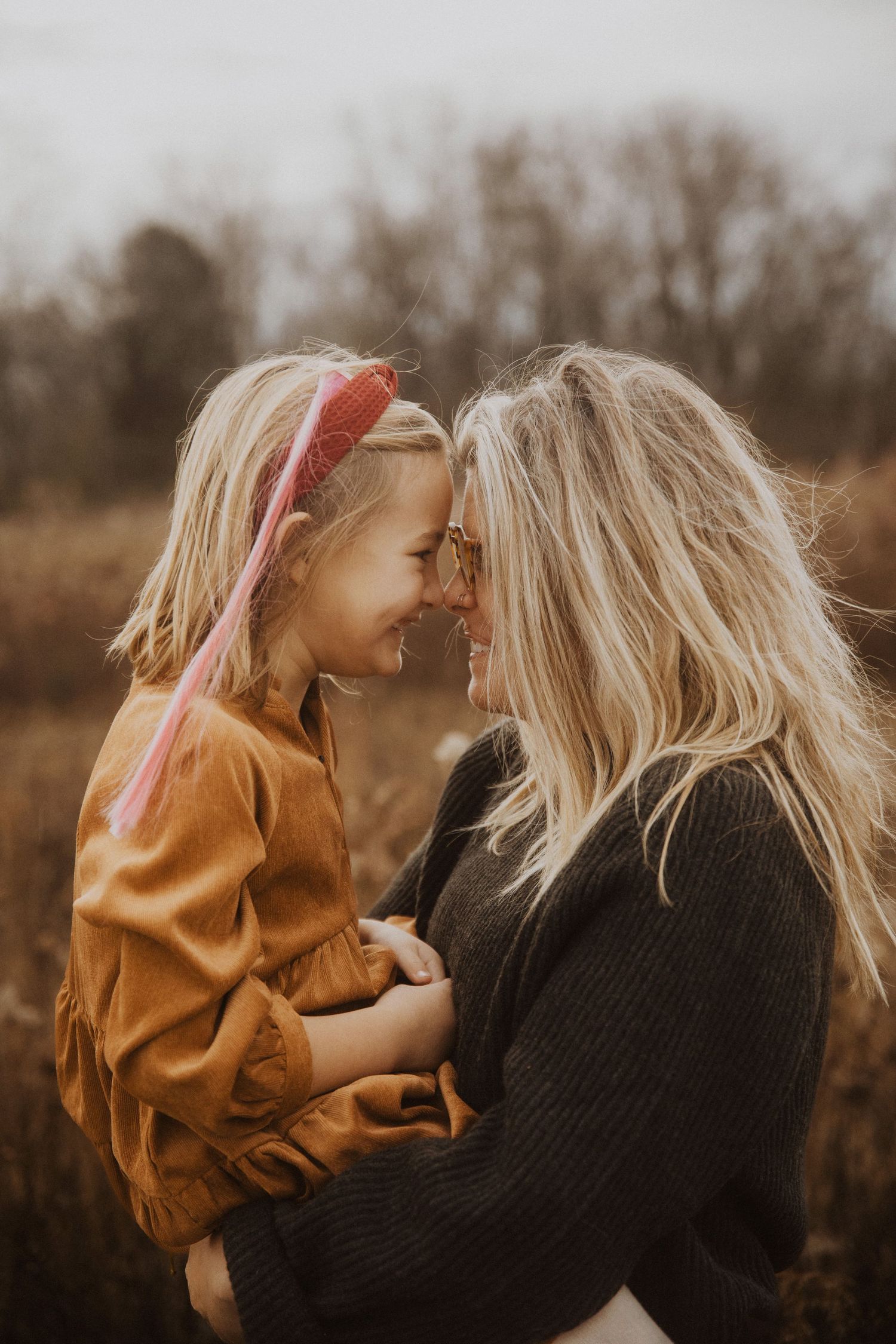 A mother holds her young blonde daughter nose-to-nose, both smiling in an autumn field.