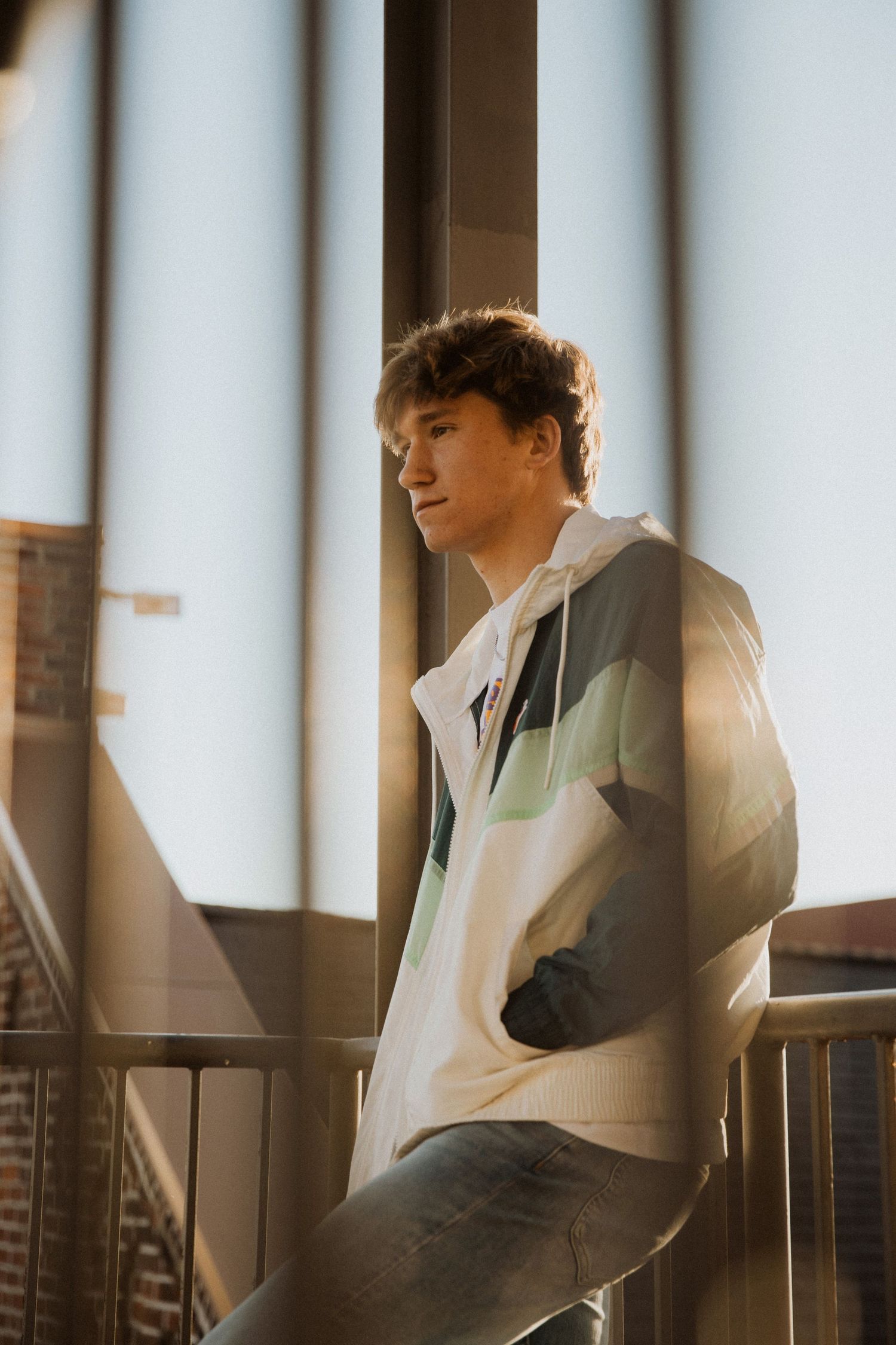 Young man in a colorful windbreaker jacket leaning on a railing near large windows with urban view.