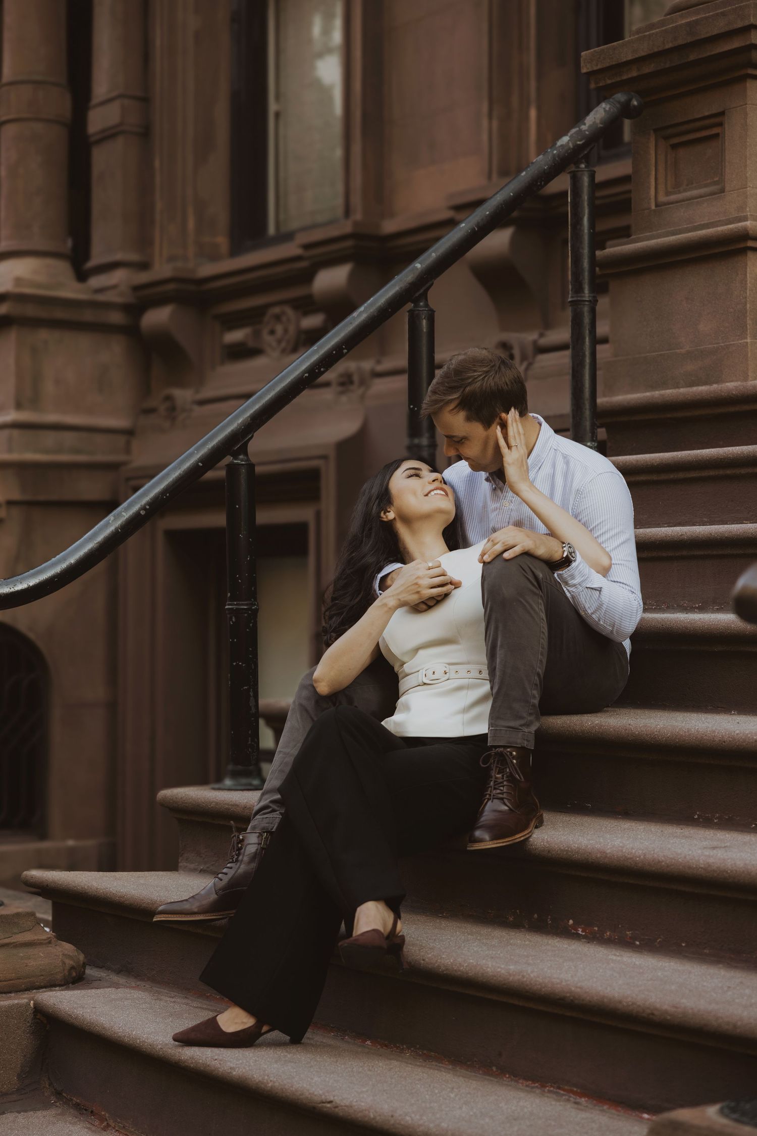 A couple shares a romantic moment on brownstone steps, the woman leaning back laughing as the man gazes at her tenderly.