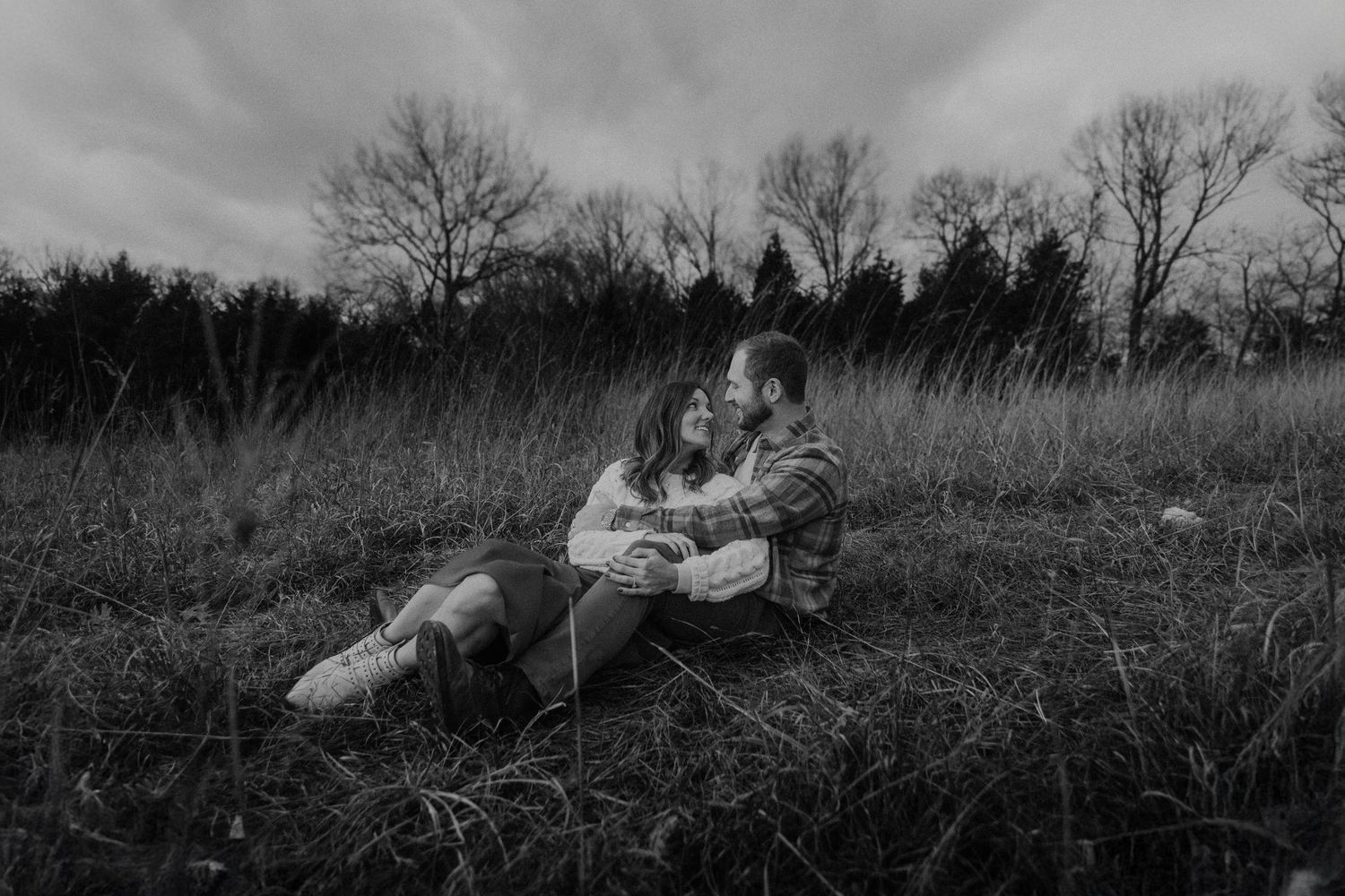 Black and white photo of a couple sitting together in a grassy field, sharing a tender moment under a cloudy sky.