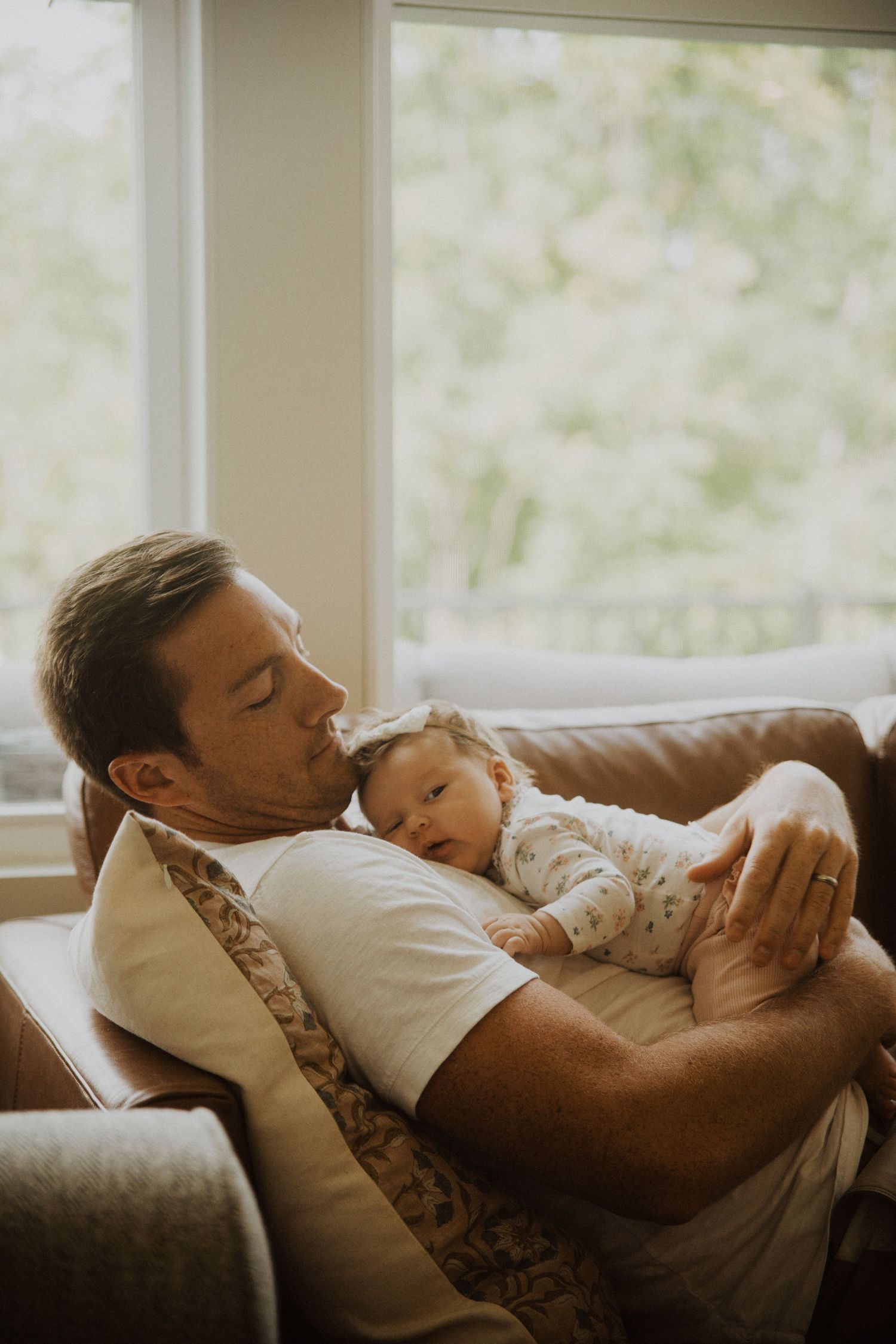 Father resting on leather sofa with sleeping newborn baby on his chest near bright window.