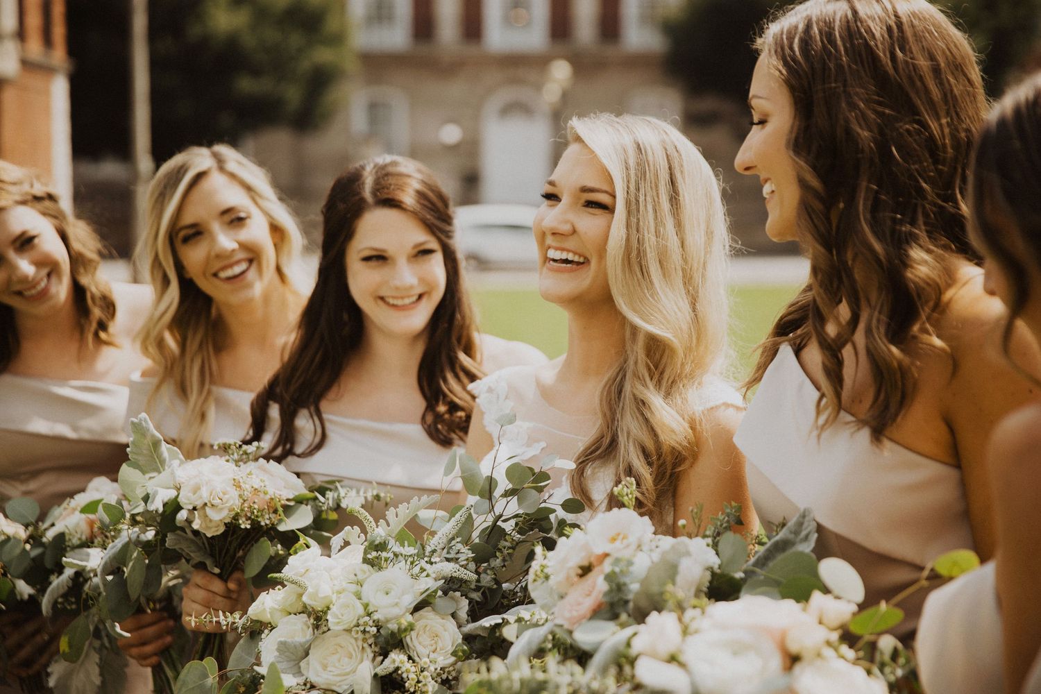Bridesmaids in strapless dresses share joyful laughter while holding elegant white and green floral bouquets.