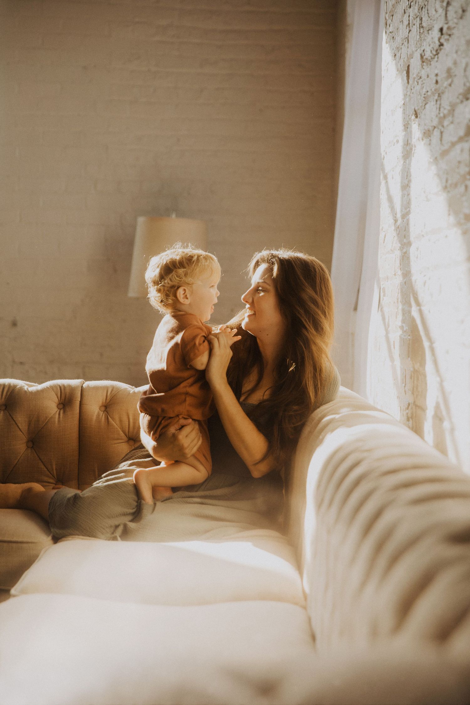 A mother and toddler share a tender moment on a tufted sofa bathed in warm natural light by a window.