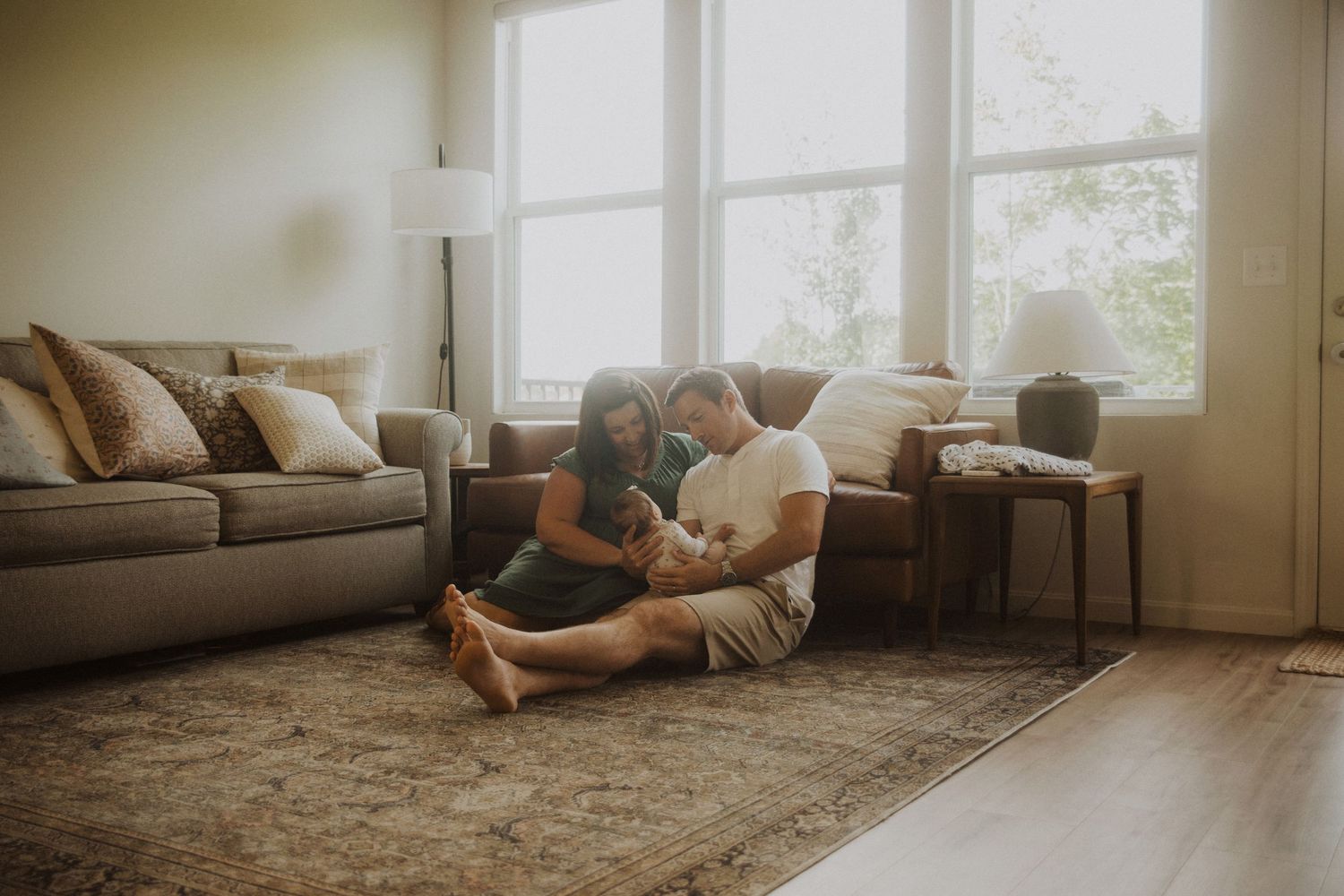 A couple sits on a rug in a bright living room, cradling their newborn baby together.