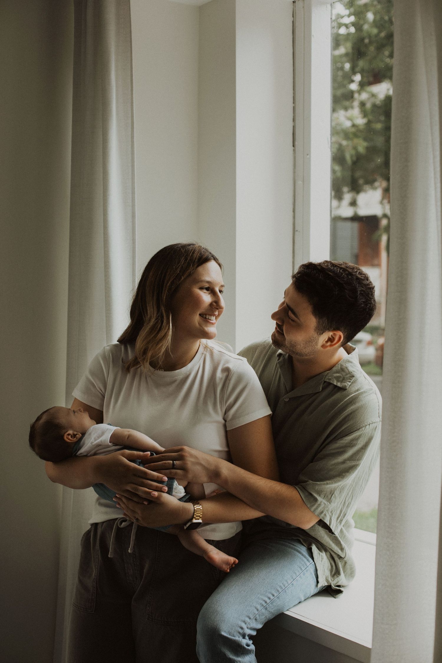 Happy young couple smiling at each other while holding their newborn baby by a bright window at home.
