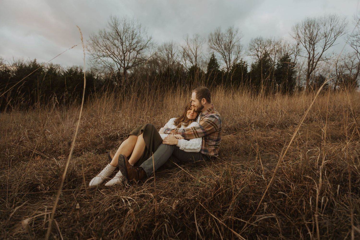 Couple sitting together in a golden field of tall grass, embracing romantically under a cloudy autumn sky.