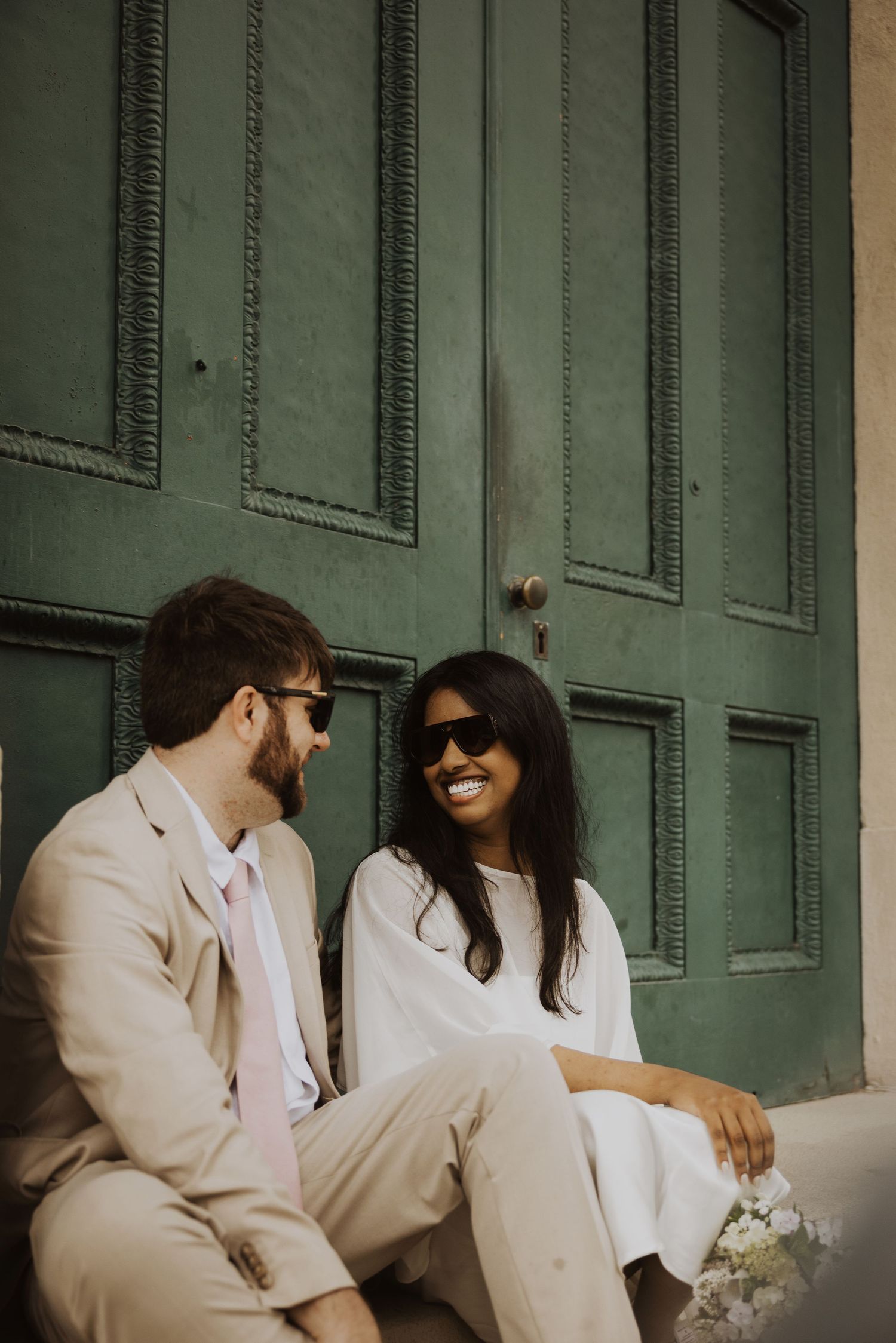Couple in beige wedding attire smiling at each other, sitting in front of a vintage green door on their wedding day.