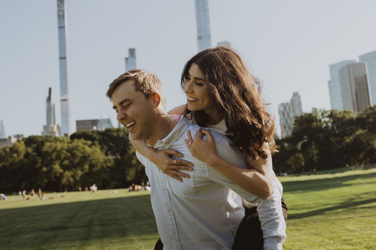 A happy couple enjoys a piggyback ride in Central Park with NYC skyline in the background on a sunny day.