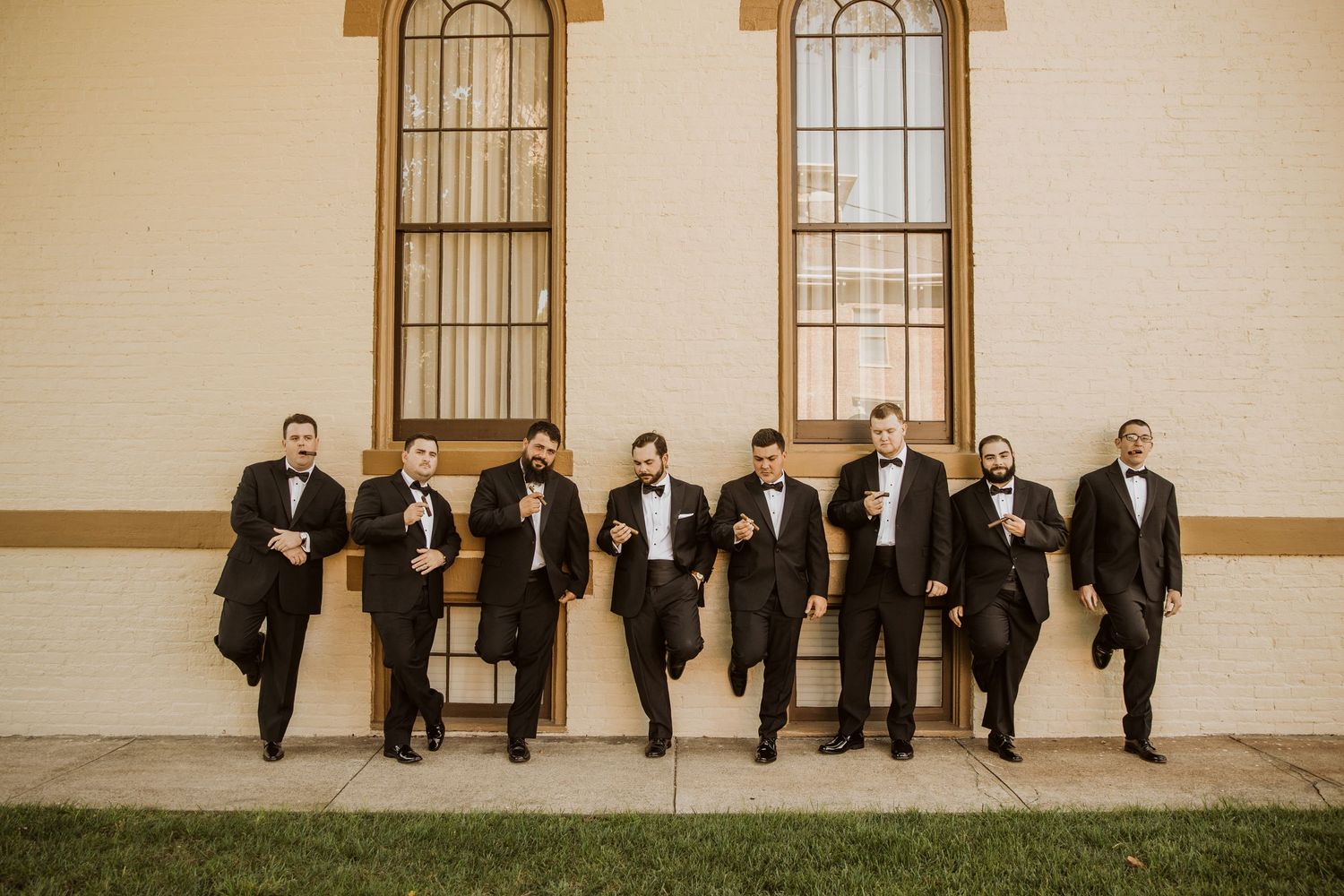 Groomsmen in black tuxedos pose together against a light colored building exterior.