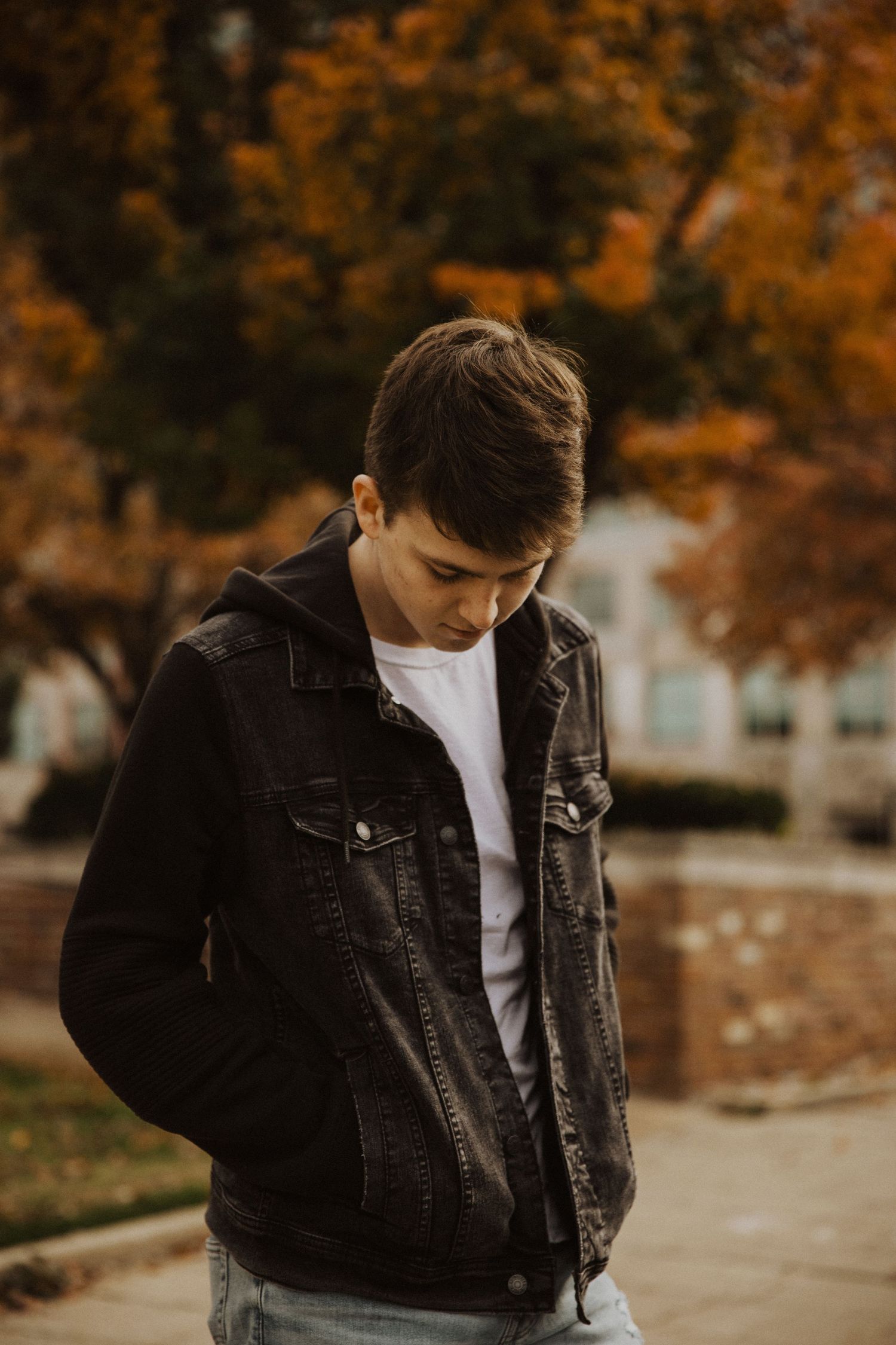 Young man in black denim jacket and white tee stands outdoors amid vibrant autumn foliage backdrop.