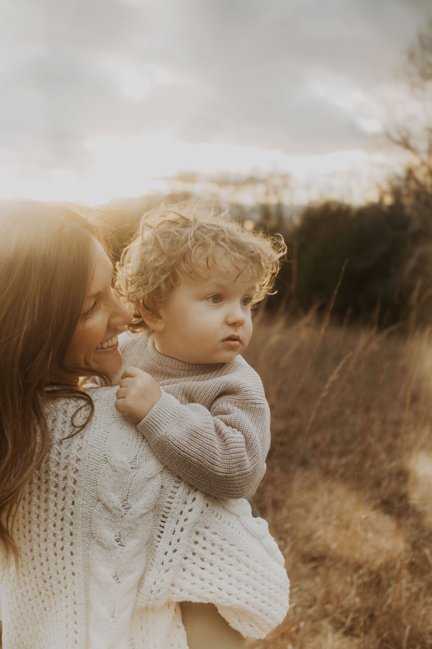 A mother holds her curly-haired toddler in a golden field at sunset, both wearing cozy knit sweaters.