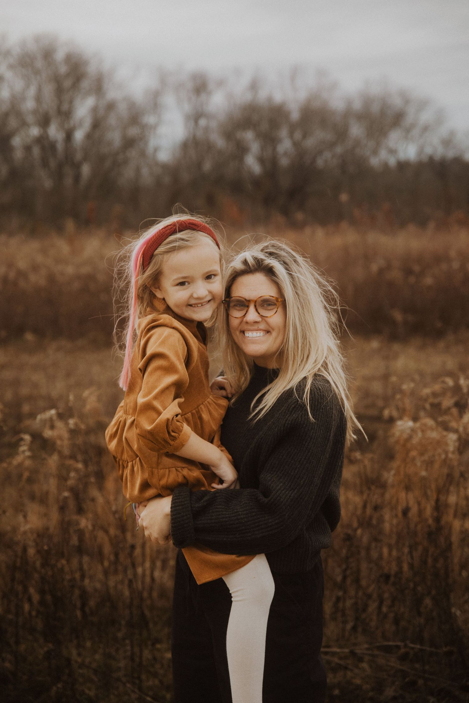 A woman with glasses holds a young girl in a mustard dress in an autumn field setting.