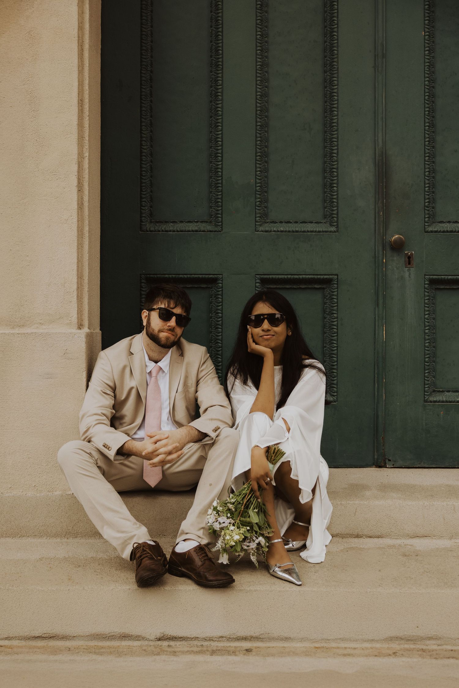 Couple in sunglasses sitting on steps before green door, man in tan suit, woman in white dress holding flowers.